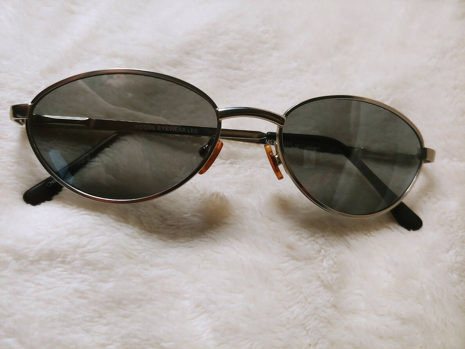 Sunglasses 桌布 壁紙 — 優雅的橢圓形太陽眼鏡，帶有深色鏡片和橙色點綴，放置在柔軟的白色毛絨表面上。
