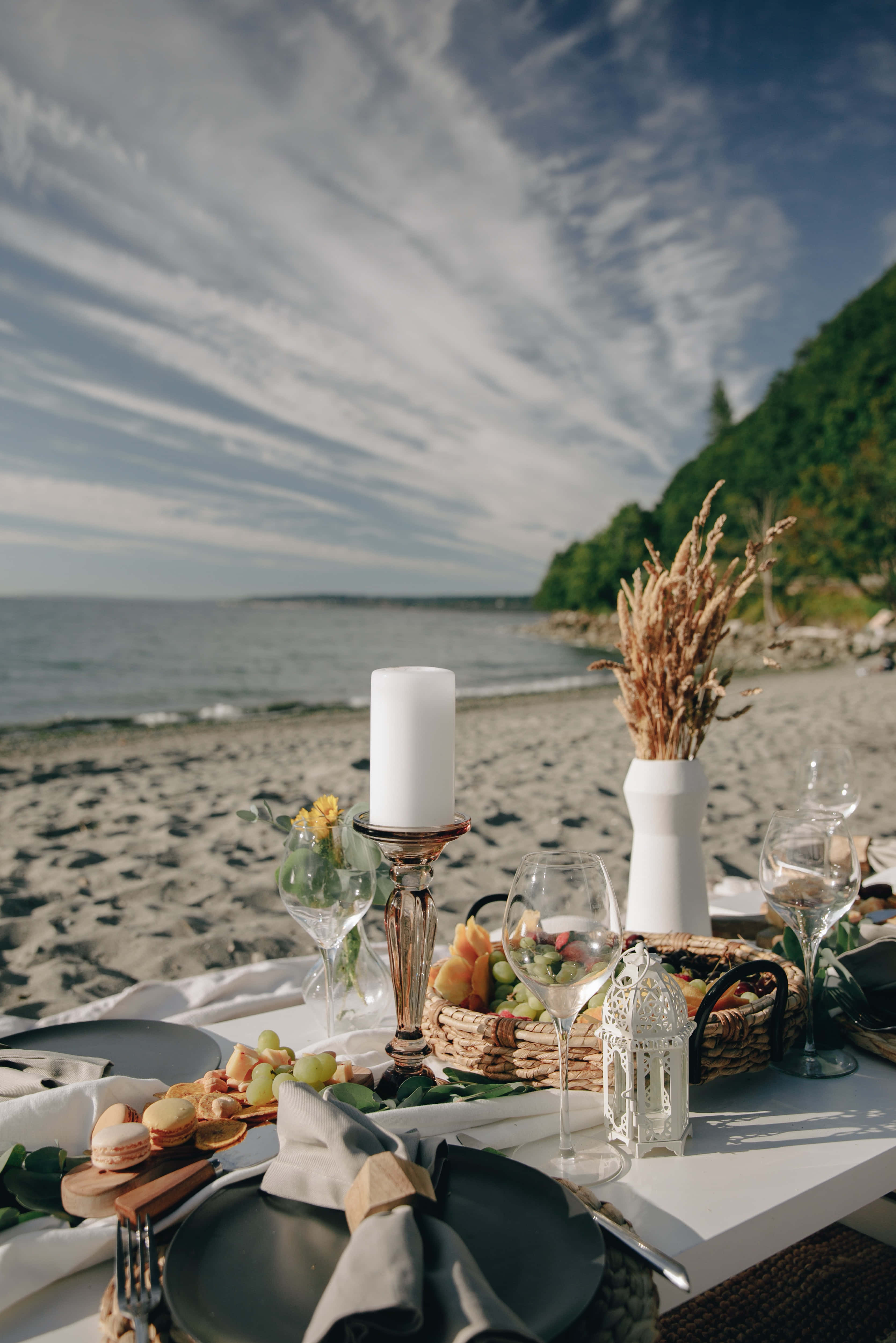 Beach Picnic 桌布 壁紙 — 優雅的海邊餐桌，擺放深色盤子、玻璃器皿和乾燥草，在明亮的天空下。