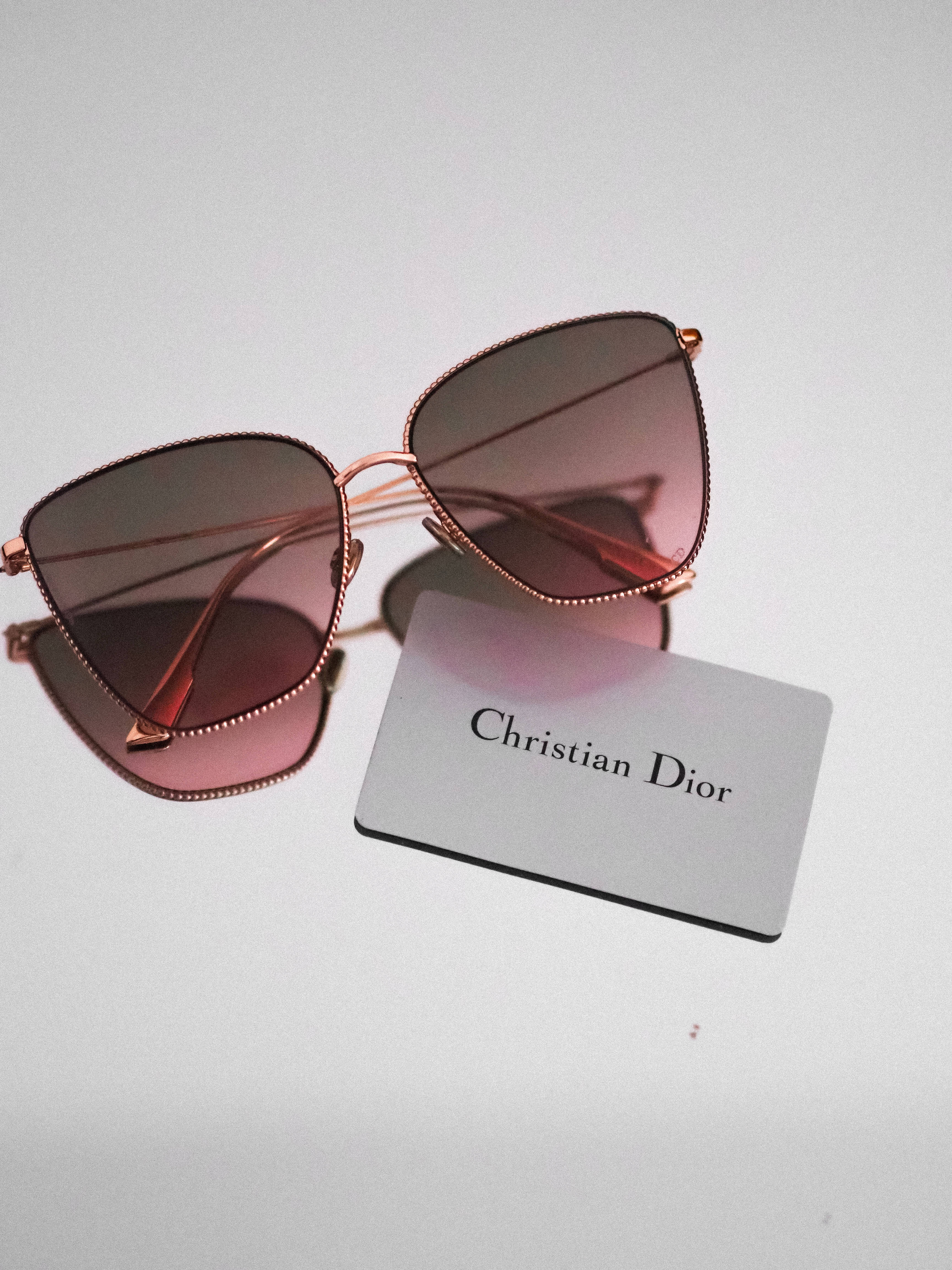 Dior 動態 桌布 壁紙 — 優雅的玫瑰金色Christian Dior太陽眼鏡，配以漸變粉紅色鏡片，放在簡約白色表面上，旁邊有一張品牌卡。