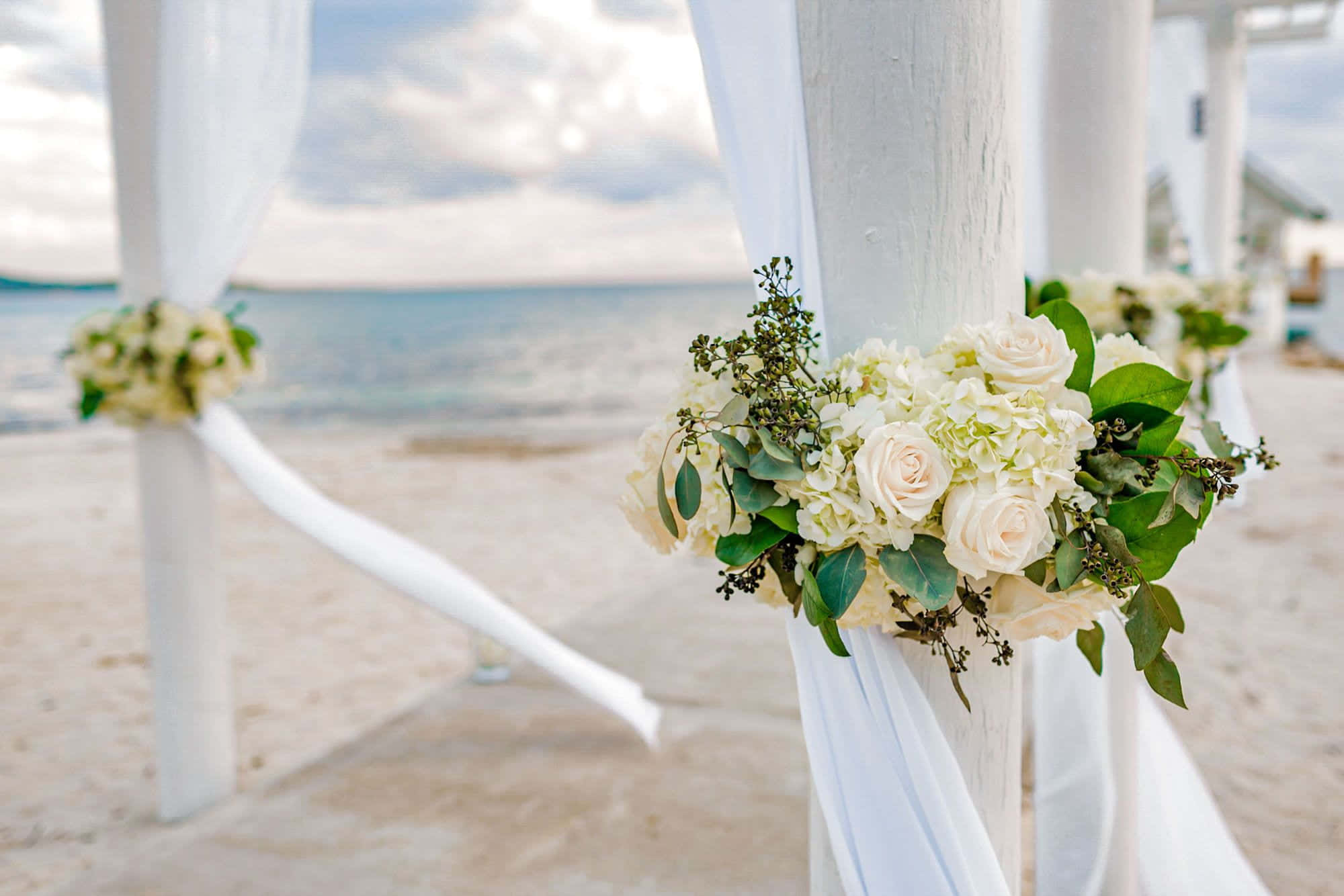 Wedding 桌布 壁紙 — 優雅的白色花卉佈置點綴著海灘婚禮拱門，搭配流動的布料，背景是平靜的海洋。