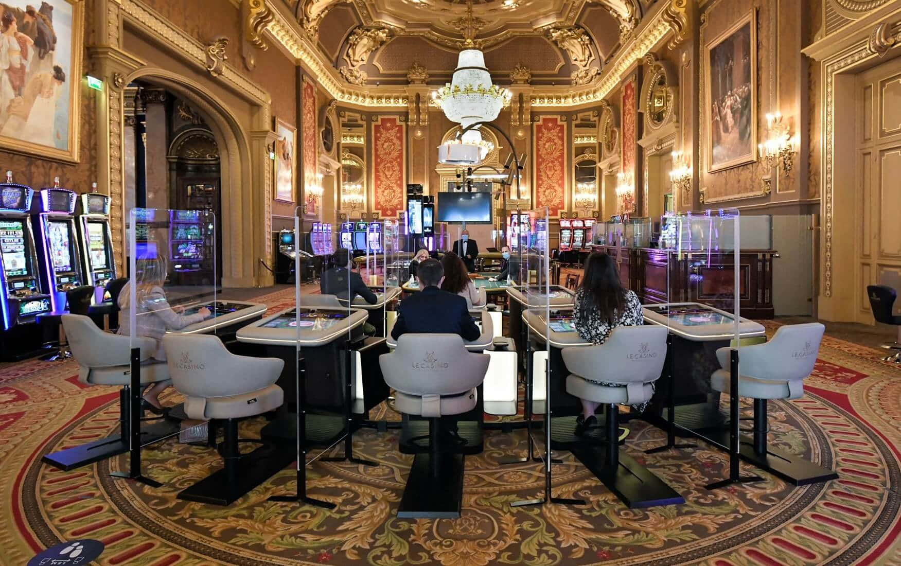 Casino 桌布 壁紙 — 優雅的賭場內部，顧客坐在遊戲桌旁，上方是華麗的水晶吊燈和金色鑲邊的牆壁。