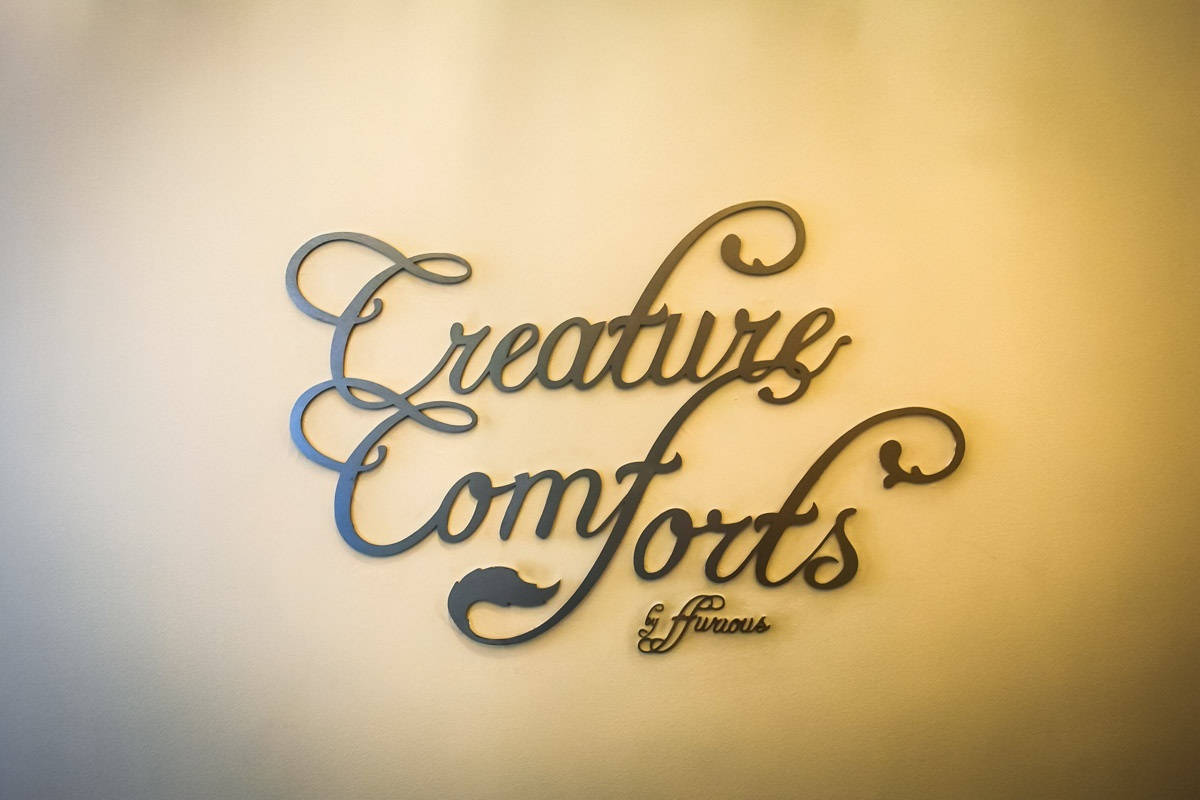 Typography 桌布 壁紙 — 優雅的金屬草書標牌，上面寫著「Creature Comforts & Fierce」，安裝在柔和照明的米色牆上。