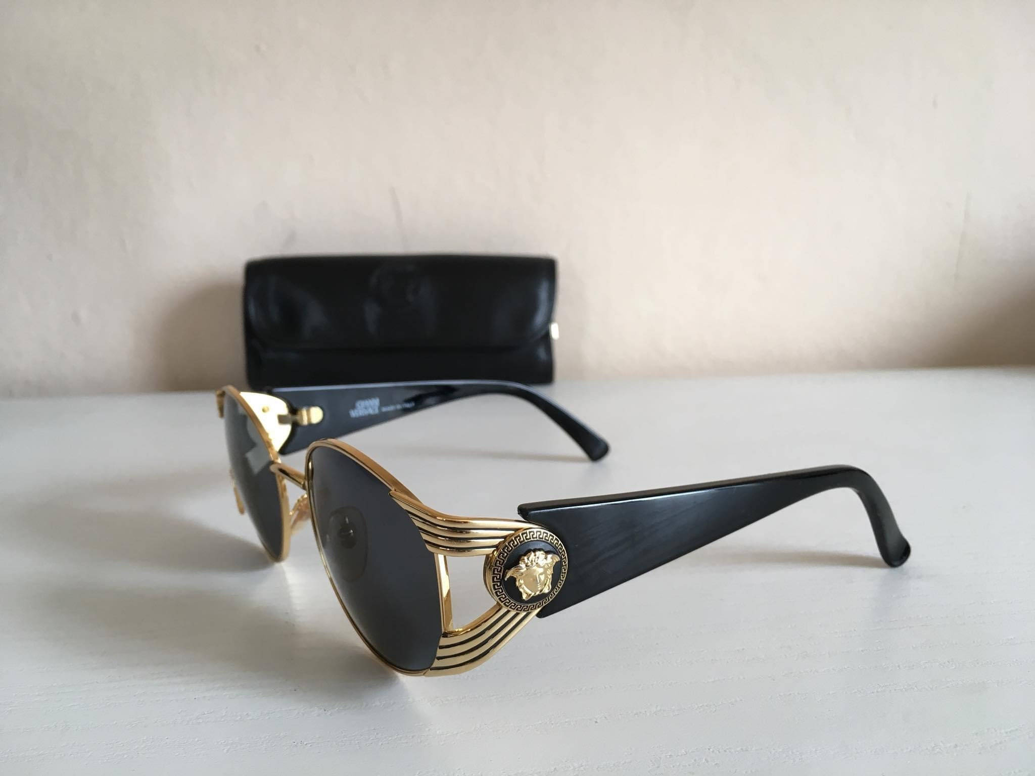 Sunglasses 桌布 壁紙 — 優雅的金色和黑色 Versace 太陽眼鏡，帶有美杜莎標誌，放置在白色表面上，旁邊有一個黑色眼鏡盒。
