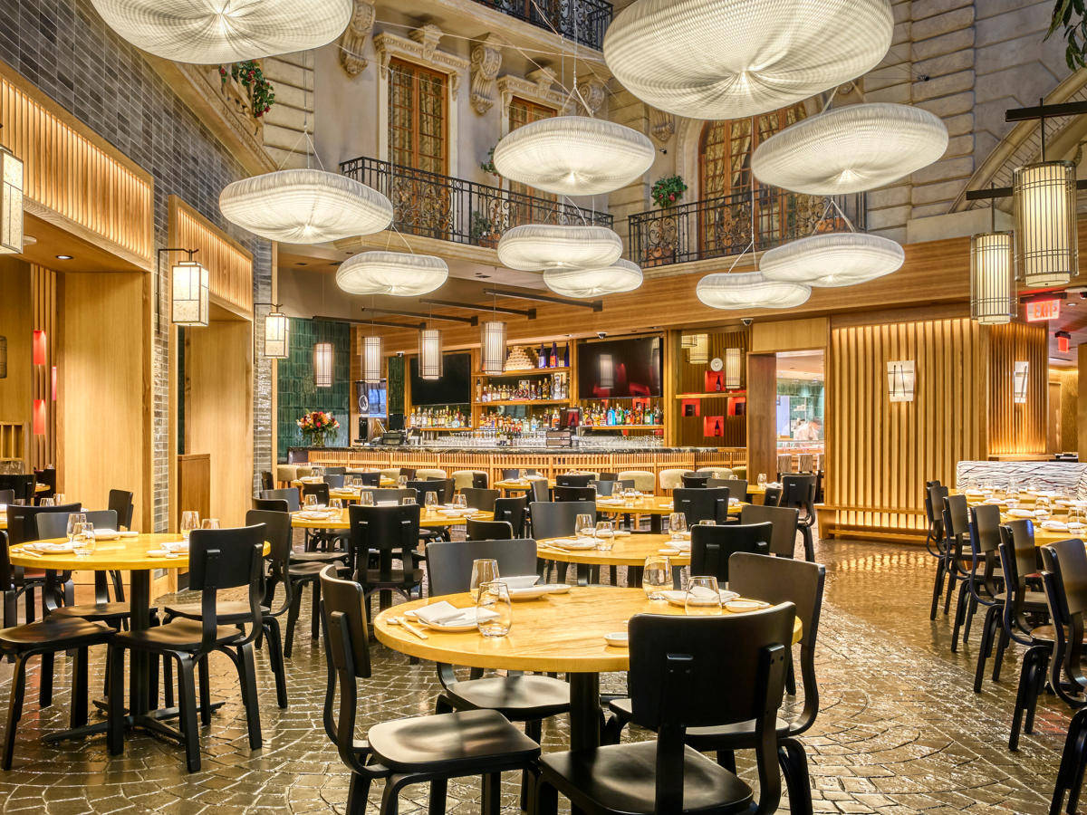 Restaurant 桌布 壁紙 — 優雅的餐廳內部，有溫暖的木桌、黑色椅子和大型編織吊燈，懸掛在高天花板下，帶有裸露的磚牆和陽台。