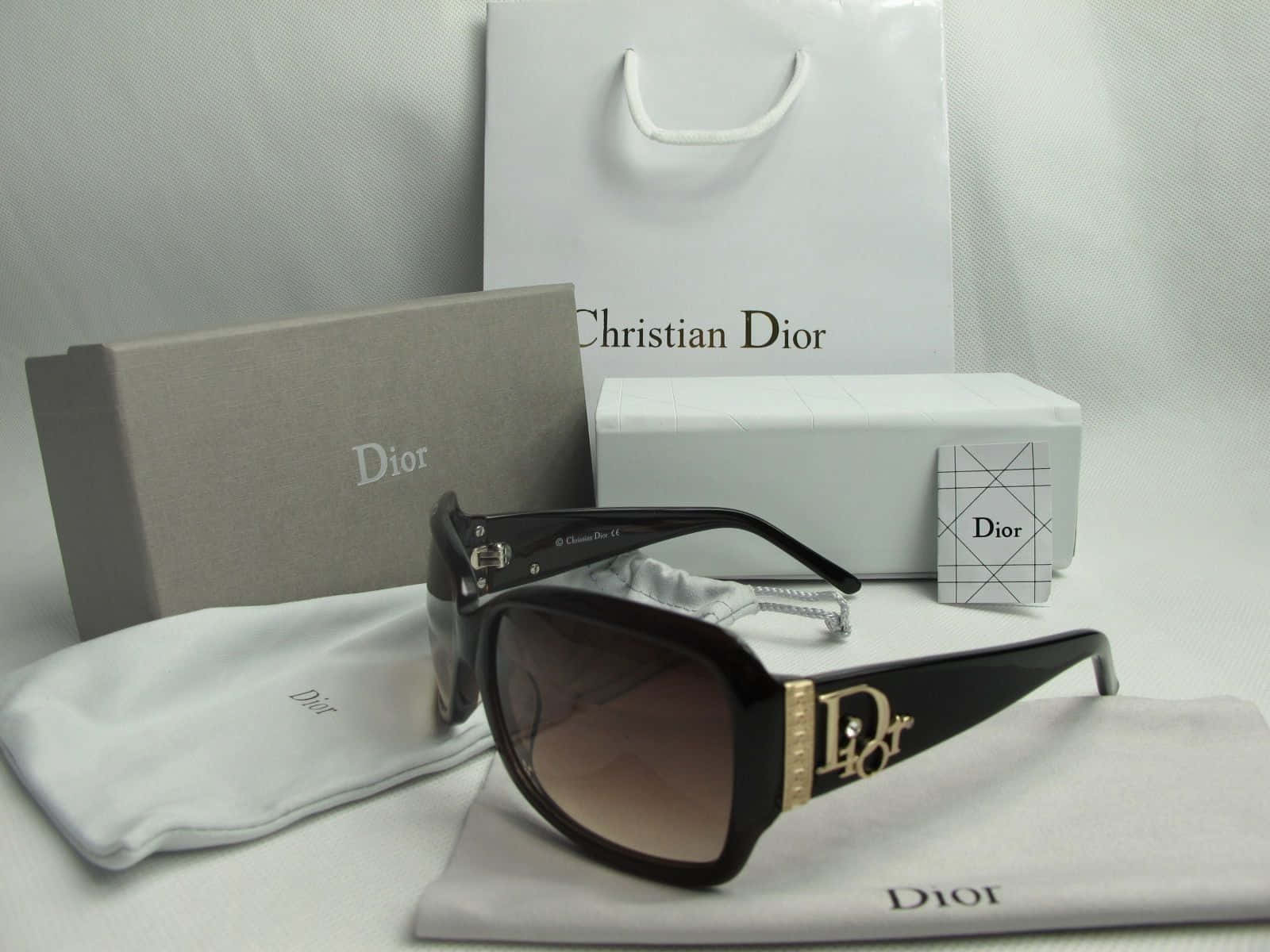 Dior 動態 桌布 壁紙 — 優雅的黑色Dior太陽眼鏡，帶有金色標誌細節，放在白色布料上，旁邊是品牌包裝，背景為中性色調。