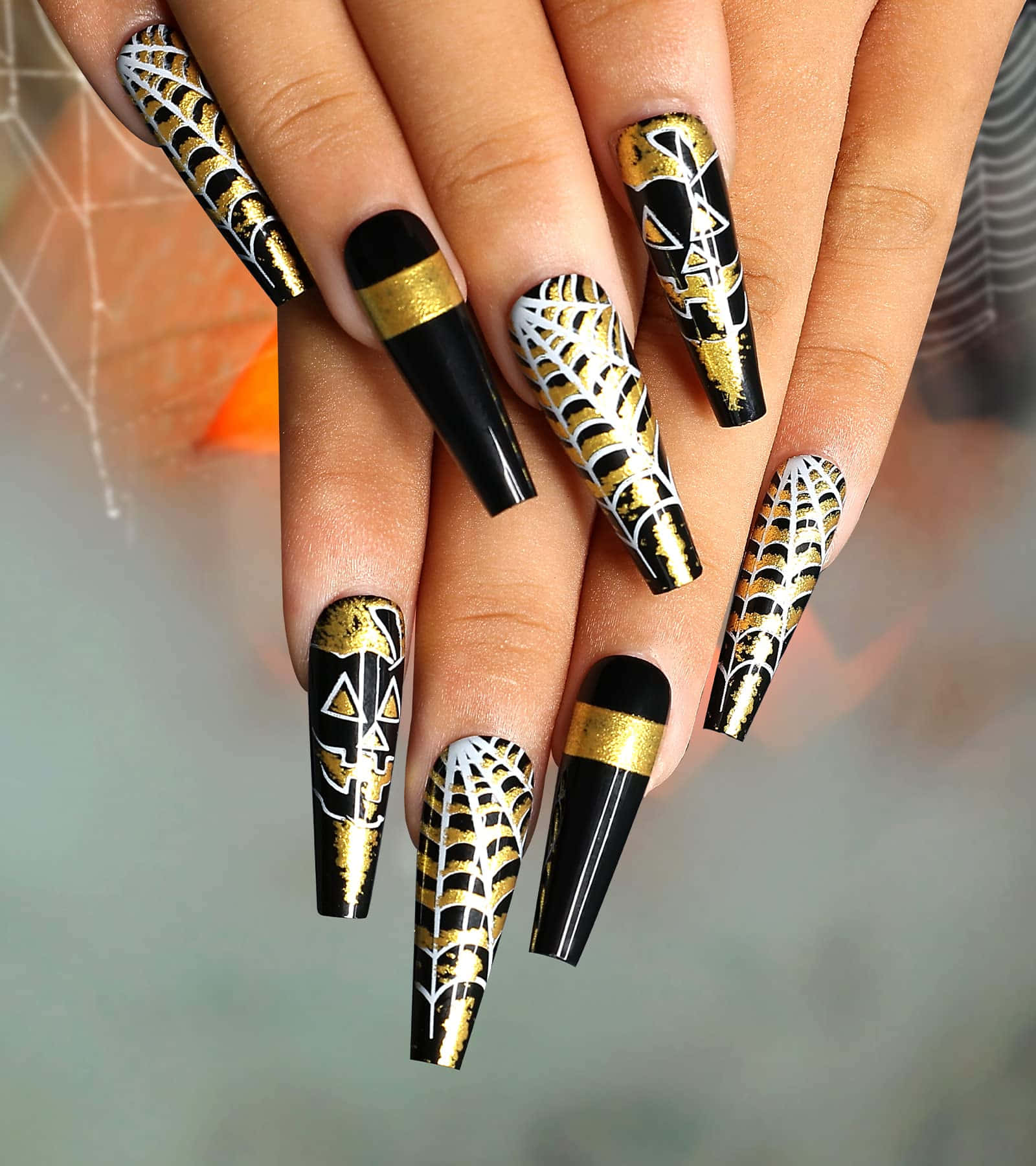Halloween Nail Art 桌布 壁紙 — 優雅的黑金蜘蛛網圖案壓克力指甲，帶有幾何圖案，手部置於模糊背景前。