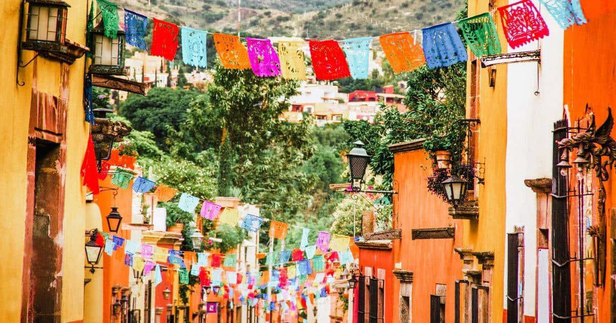 墨西哥 桌布 壁紙 — 充滿活力的 papel picado 橫幅在陽光明媚的墨西哥街道上，在暖色調的殖民建築之間飄揚。