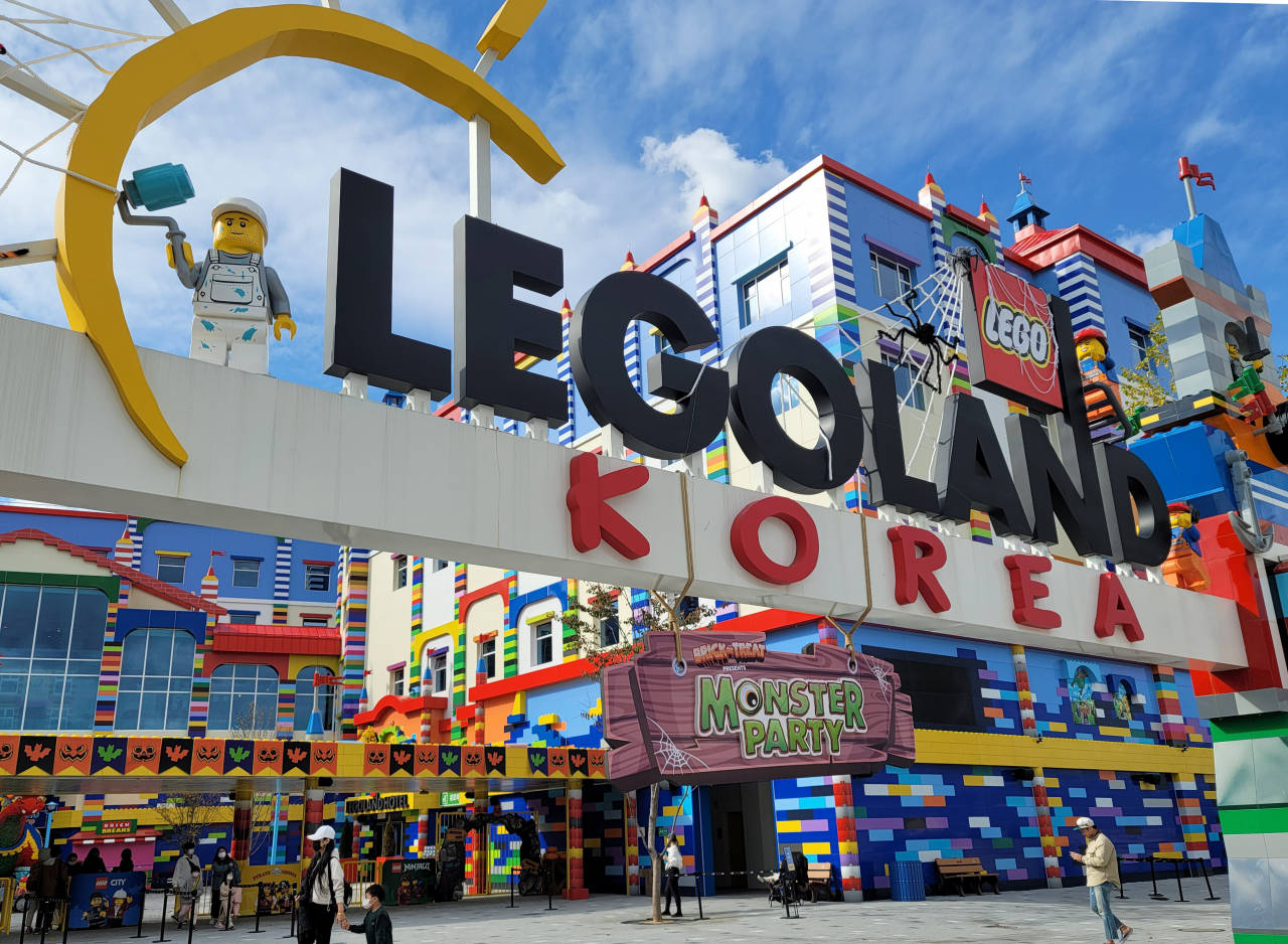 小孩動圖 桌布 壁紙 — 充滿活力的LEGO主題遊樂園入口，色彩繽紛的積木建築和一個巨大的LEGO人偶在明亮的藍天下拿著畫筆。