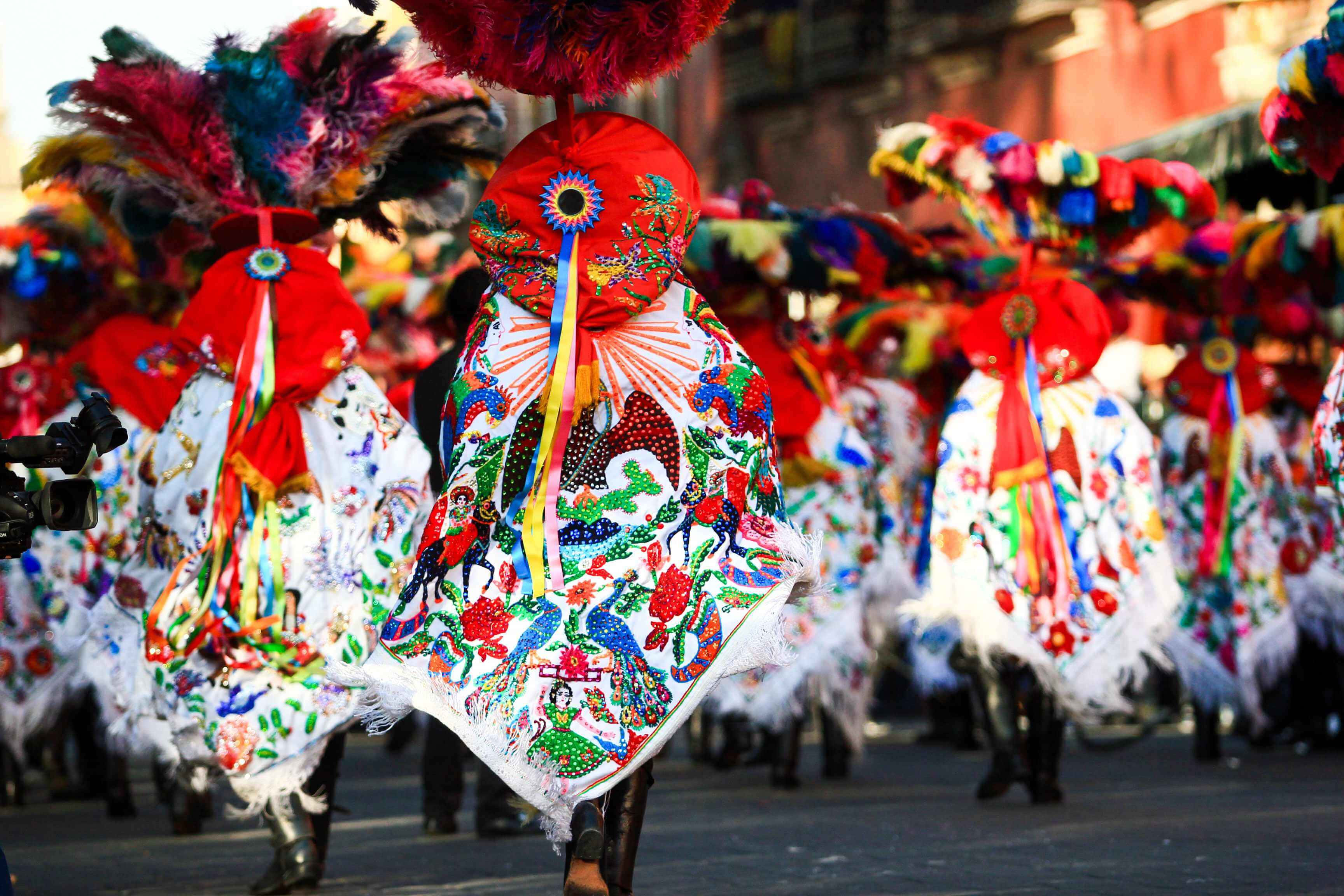 Mexican 桌布 壁紙 — 充滿活力的傳統舞者身穿精緻的羽毛服裝，在陽光普照的街頭遊行中充滿活力地表演。