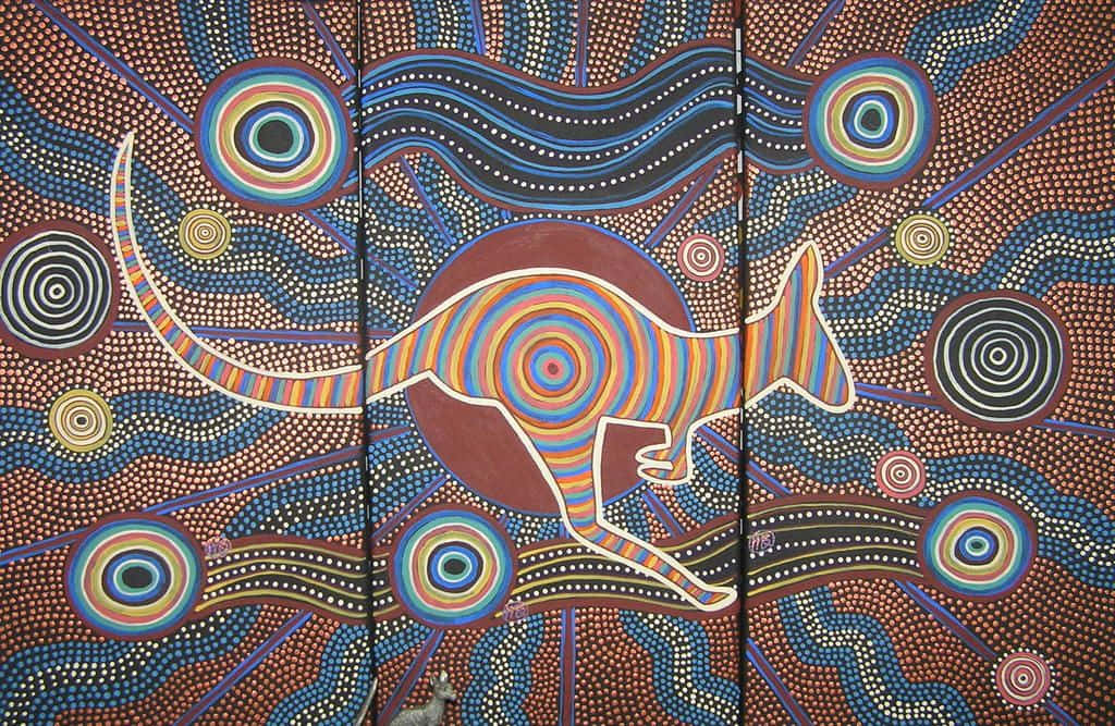 Indigenous 桌布 壁紙 — 充滿活力的原住民點畫描繪了一隻色彩繽紛的袋鼠在豐富的棕色背景上，在漩渦圖案和同心圓之間跳躍。