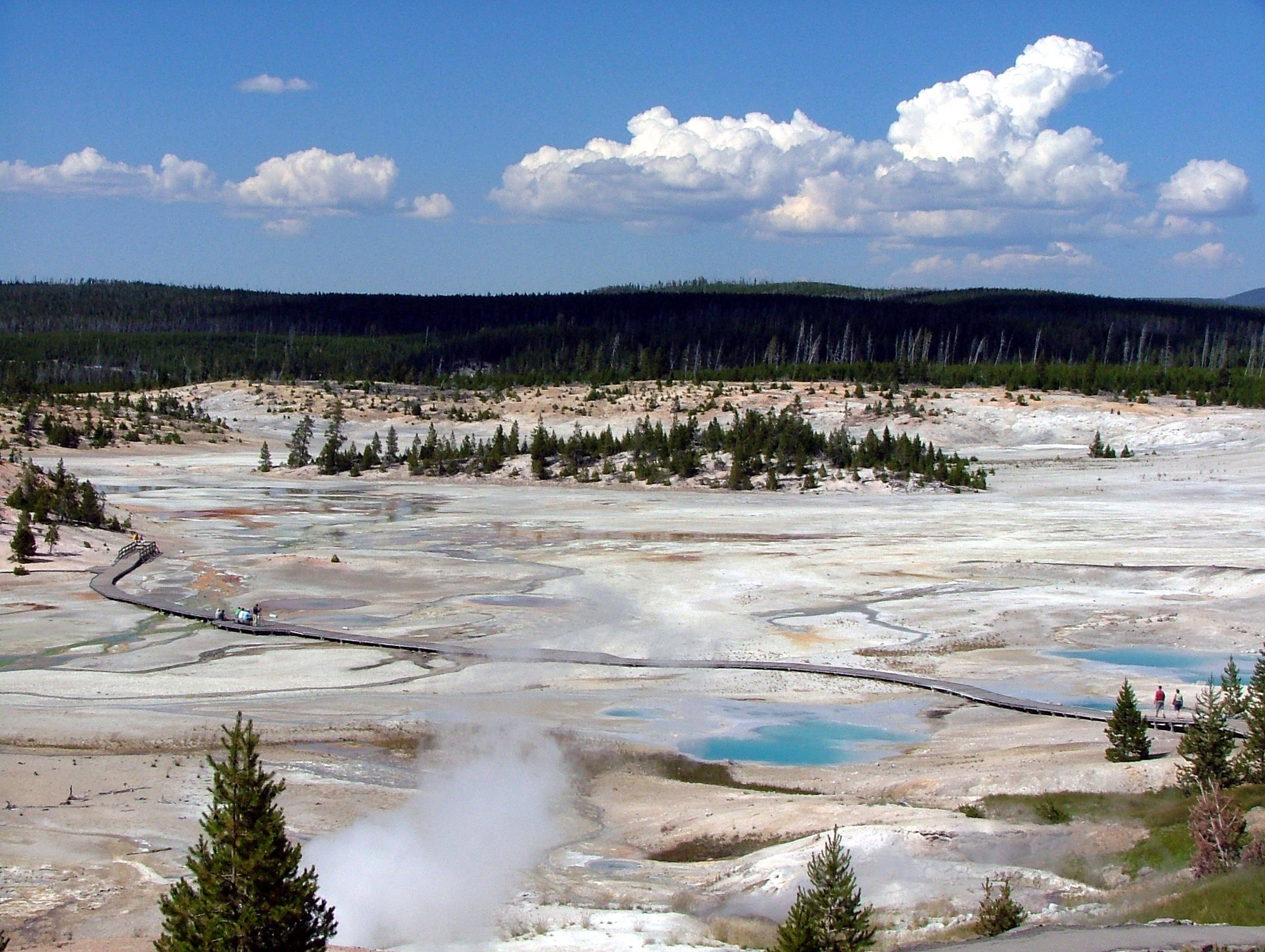 Yellowstone 桌布 壁紙 — 充滿活力的地熱景觀，有冒煙的池子和色彩繽紛的礦物沉積物，在明亮的藍天下有蓬鬆的雲朵。