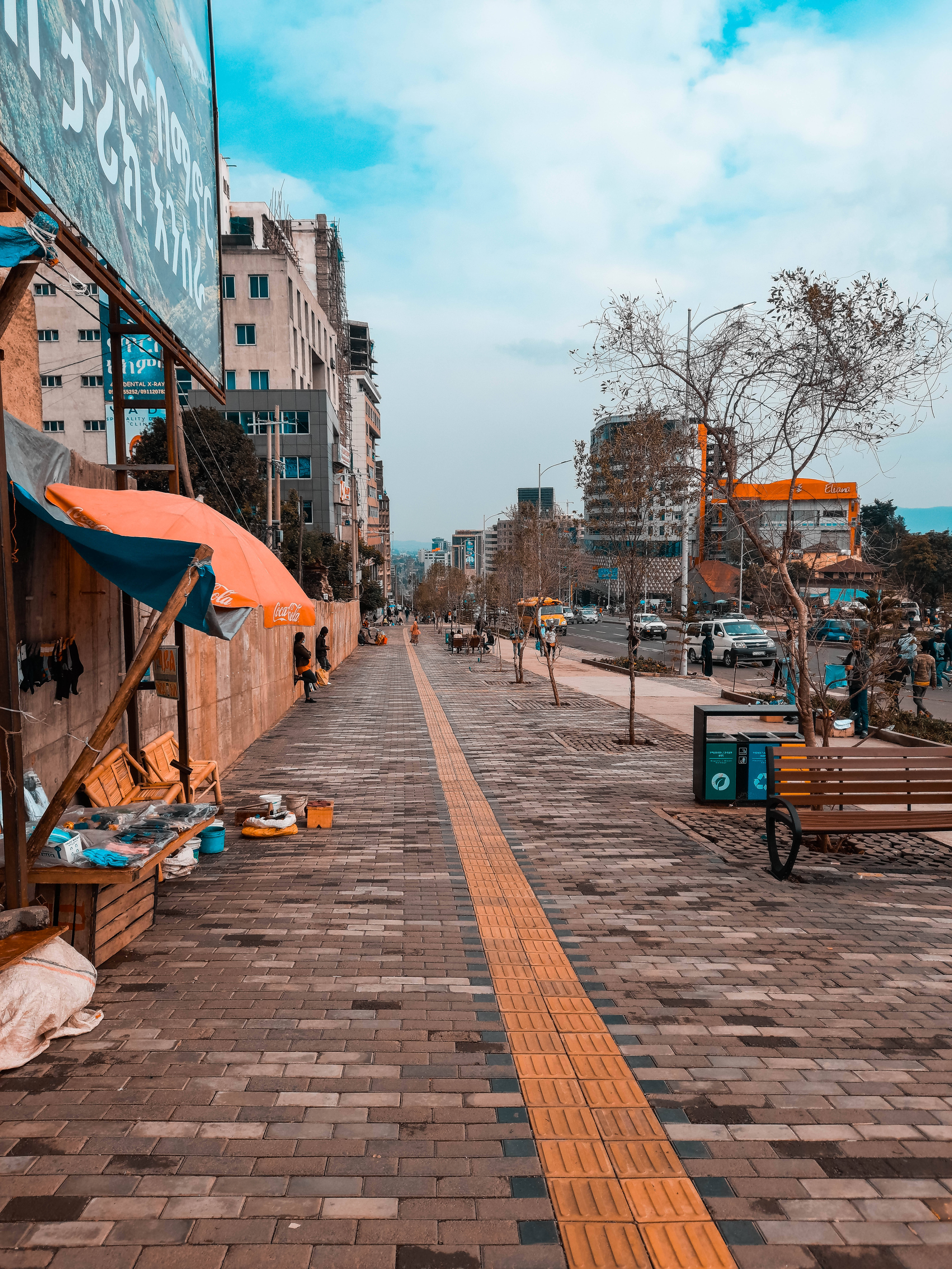 Ethiopia 桌布 壁紙 — 充滿活力的城市人行道，鋪有磚塊路面、橙色觸覺引道、攤販攤位和光禿禿的樹木，天空部分多雲。
