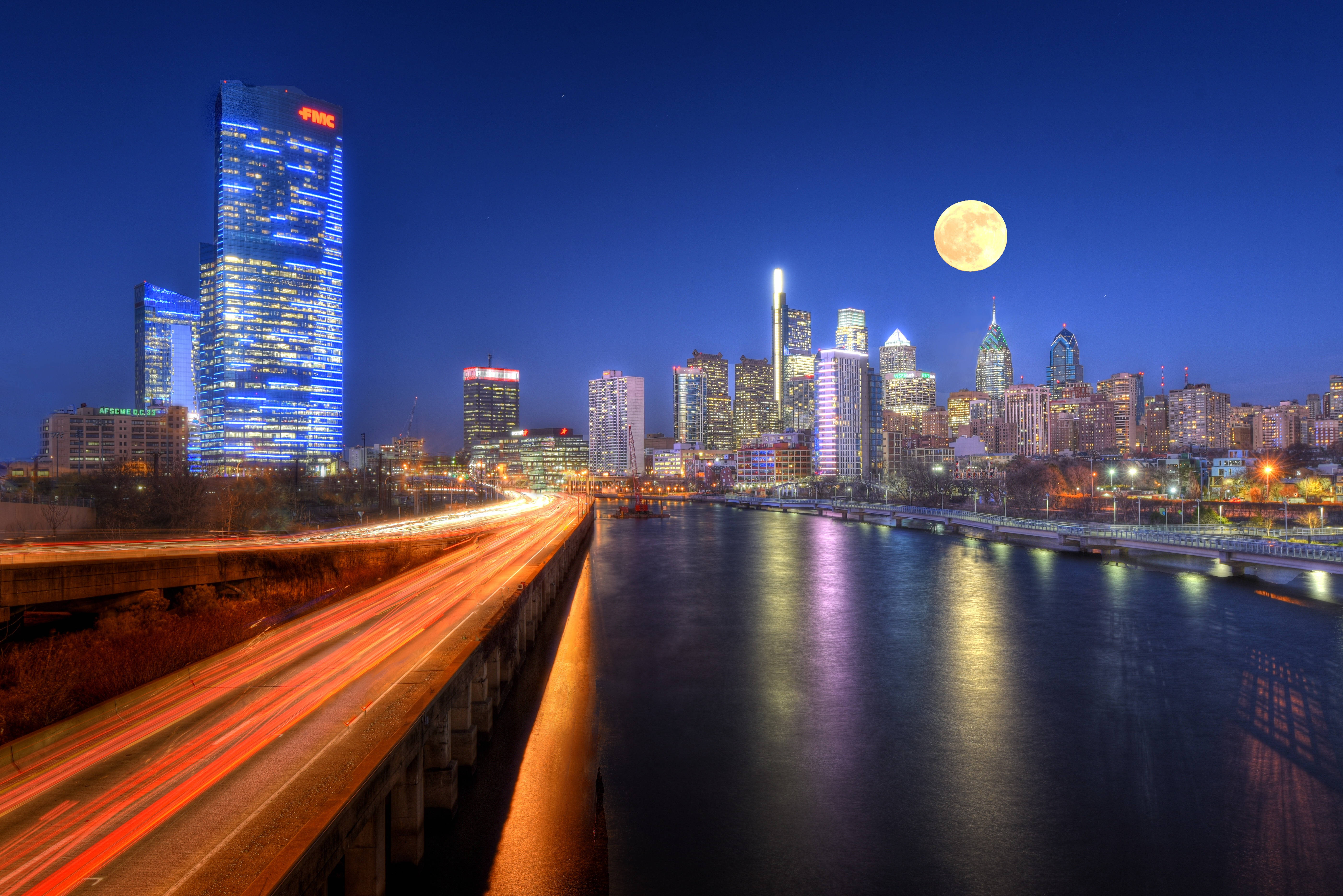 Philadelphia 桌布 壁紙 — 充滿活力的城市夜景，滿月在摩天大樓上方閃耀，繁忙高速公路的光跡在反射的河流旁。