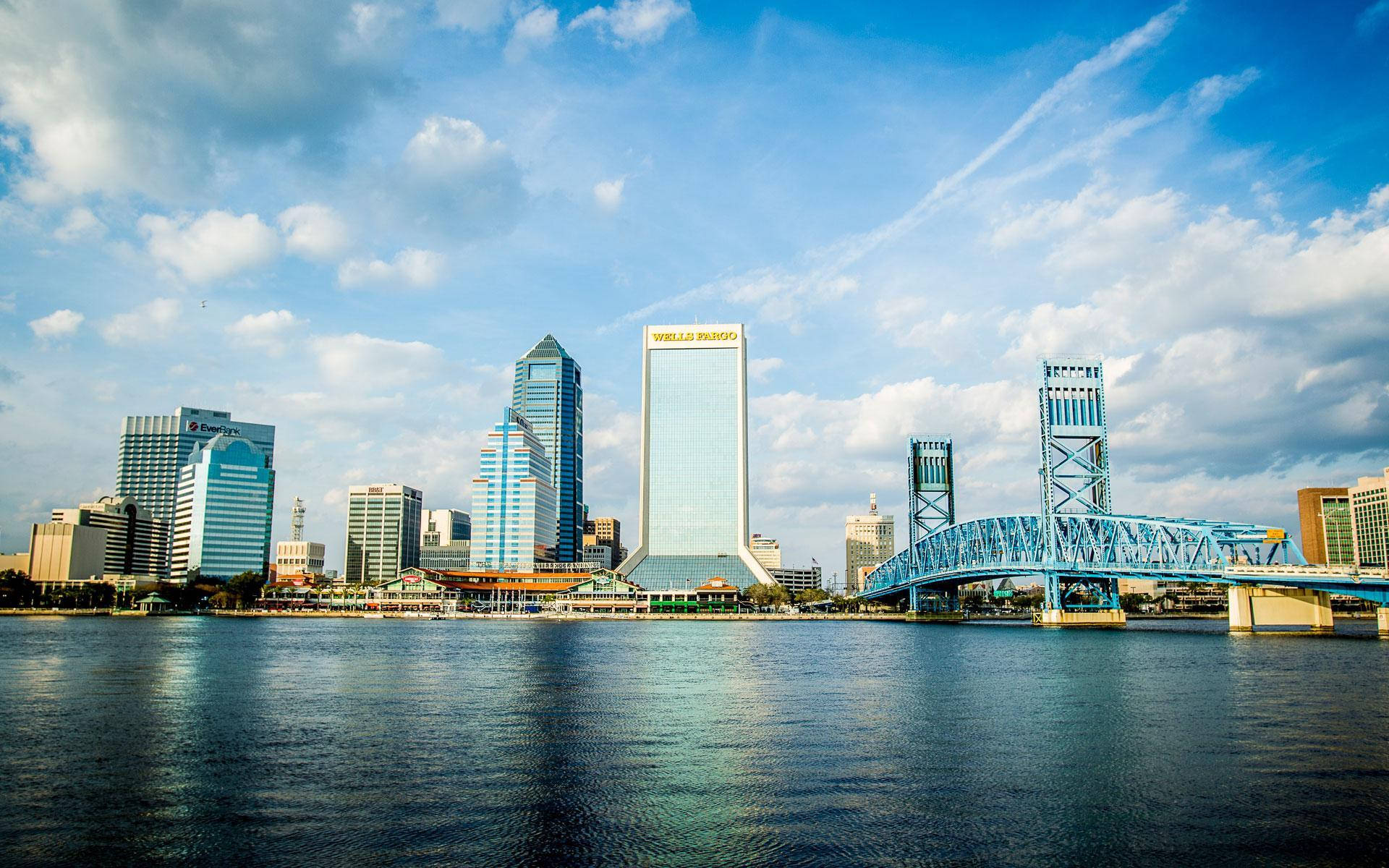 Jacksonville 桌布 壁紙 — 充滿活力的城市天際線，現代摩天大樓和一座藍色橋樑橫跨平靜的河流，天空部分多雲。