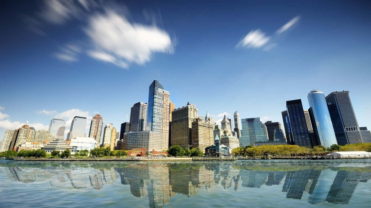 720p Ny City 桌布 壁紙 — 充滿活力的城市天際線，高聳的摩天大樓倒映在平靜的水面上，天空湛藍，雲彩斑駁。