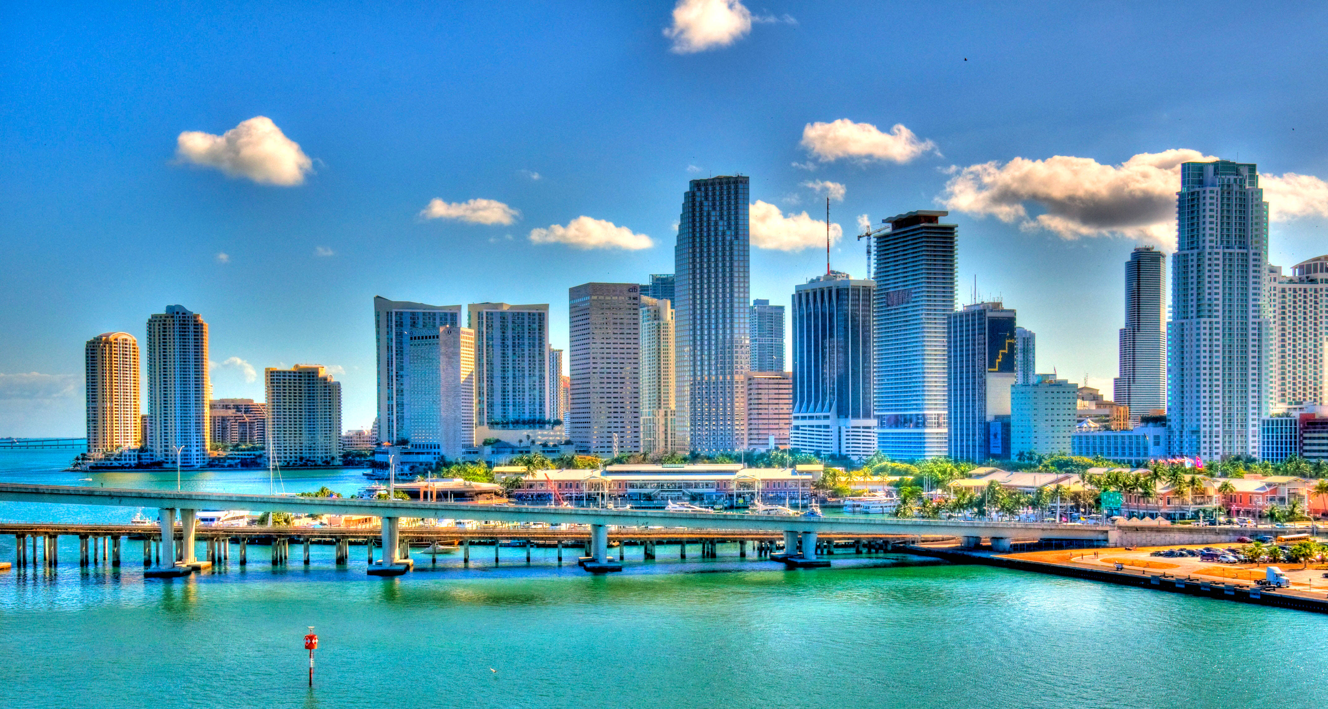Miami 桌布 壁紙 — 充滿活力的城市天際線，高聳的摩天大樓倒映在碧綠色的海水中，上方是晴朗的藍天和蓬鬆的白雲。