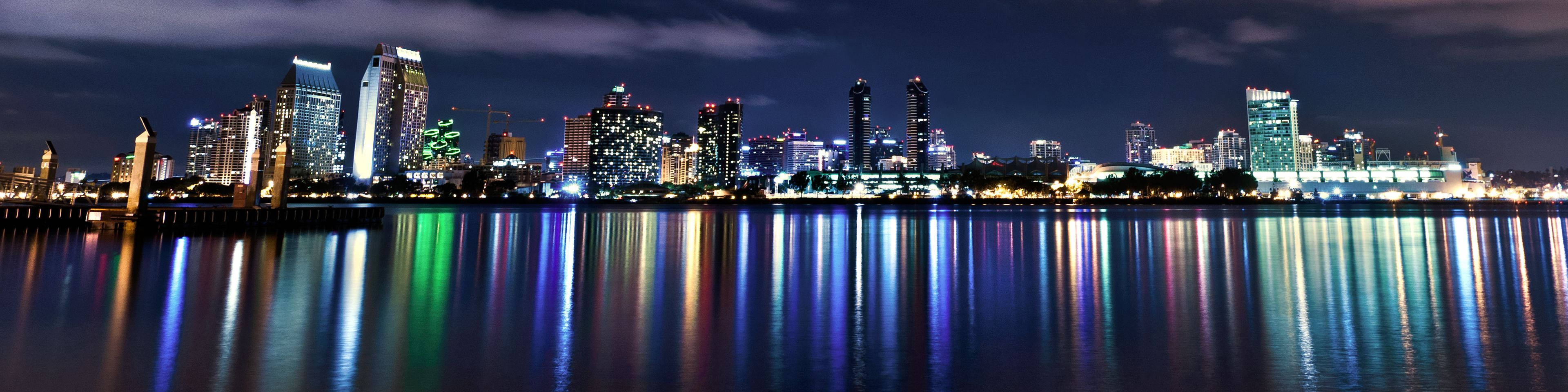 3 Monitor 桌布 壁紙 — 充滿活力的城市天際線全景夜景，倒映在平靜的水面上，色彩繽紛的燈光在水面閃爍。