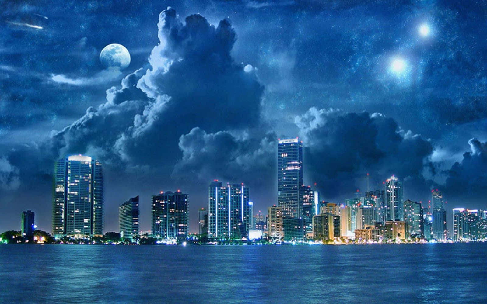 風景 桌布 壁紙 — 充滿活力的城市天際線在夜晚倒映在平靜的水面上，月光下天空中有戲劇性的雲彩和星星。