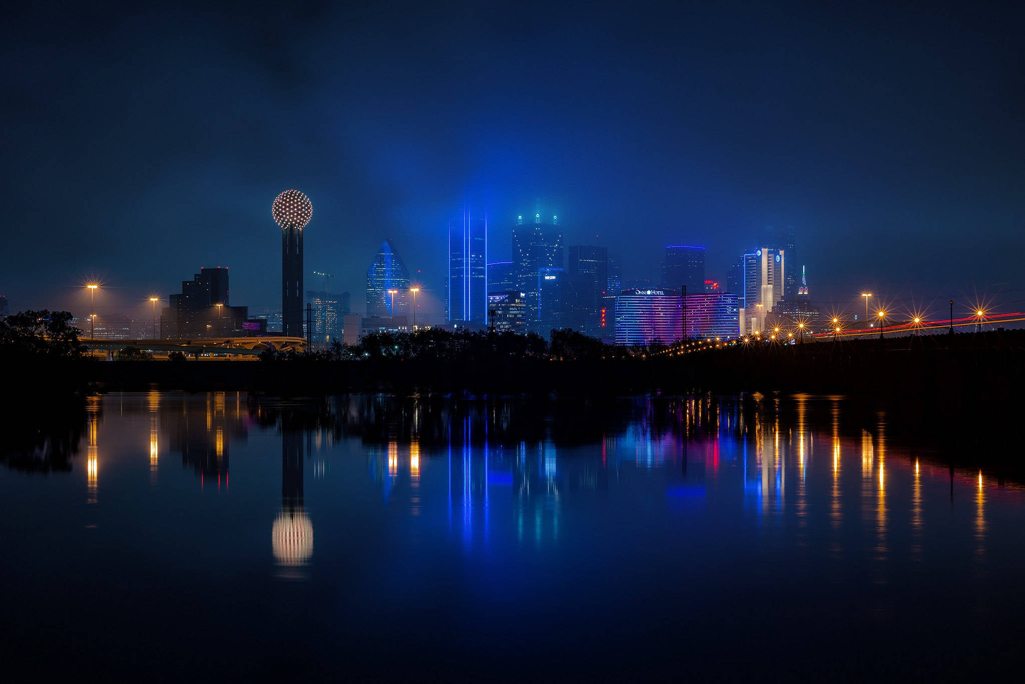 城市 桌布 壁紙 — 充滿活力的城市天際線在夜晚倒映在平靜的水面上，伴隨著五彩繽紛的燈光和發光的球形塔。