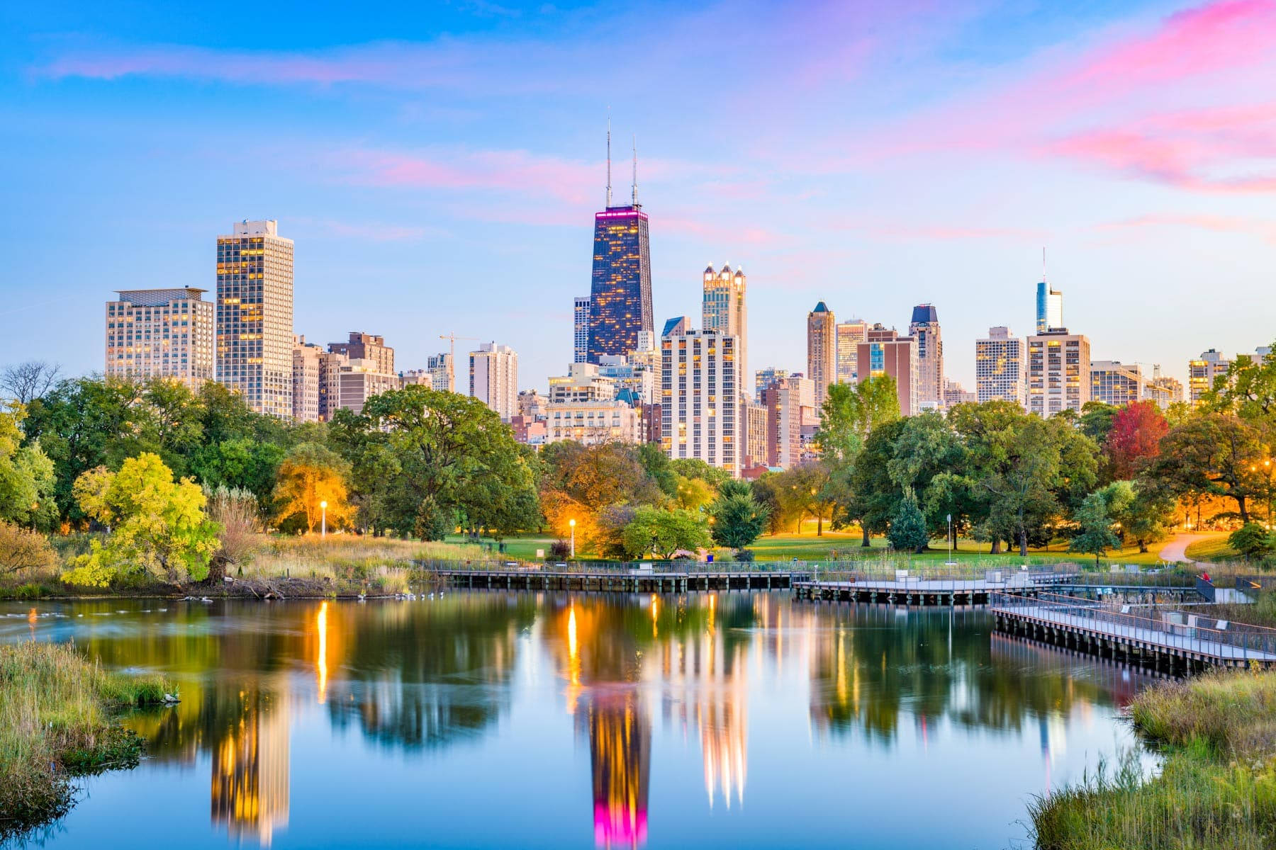 Illinois 桌布 壁紙 — 充滿活力的城市天際線在日落時分倒映在平靜的湖面上，周圍環繞著茂密的綠色公園樹木。