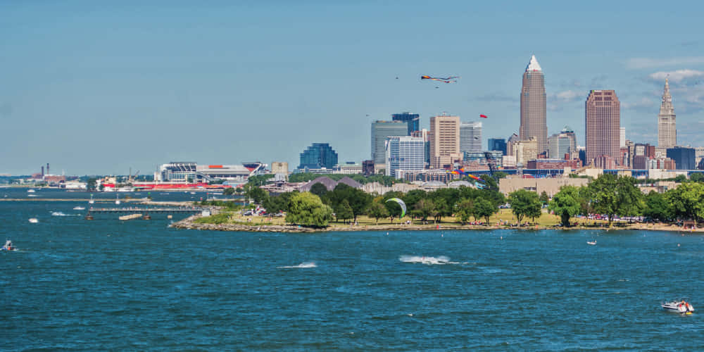 Lake Erie 桌布 壁紙 — 充滿活力的城市天際線在樹木成行的海濱後方升起，有船隻和懸掛式滑翔機在晴朗天空下飛越藍色水面。