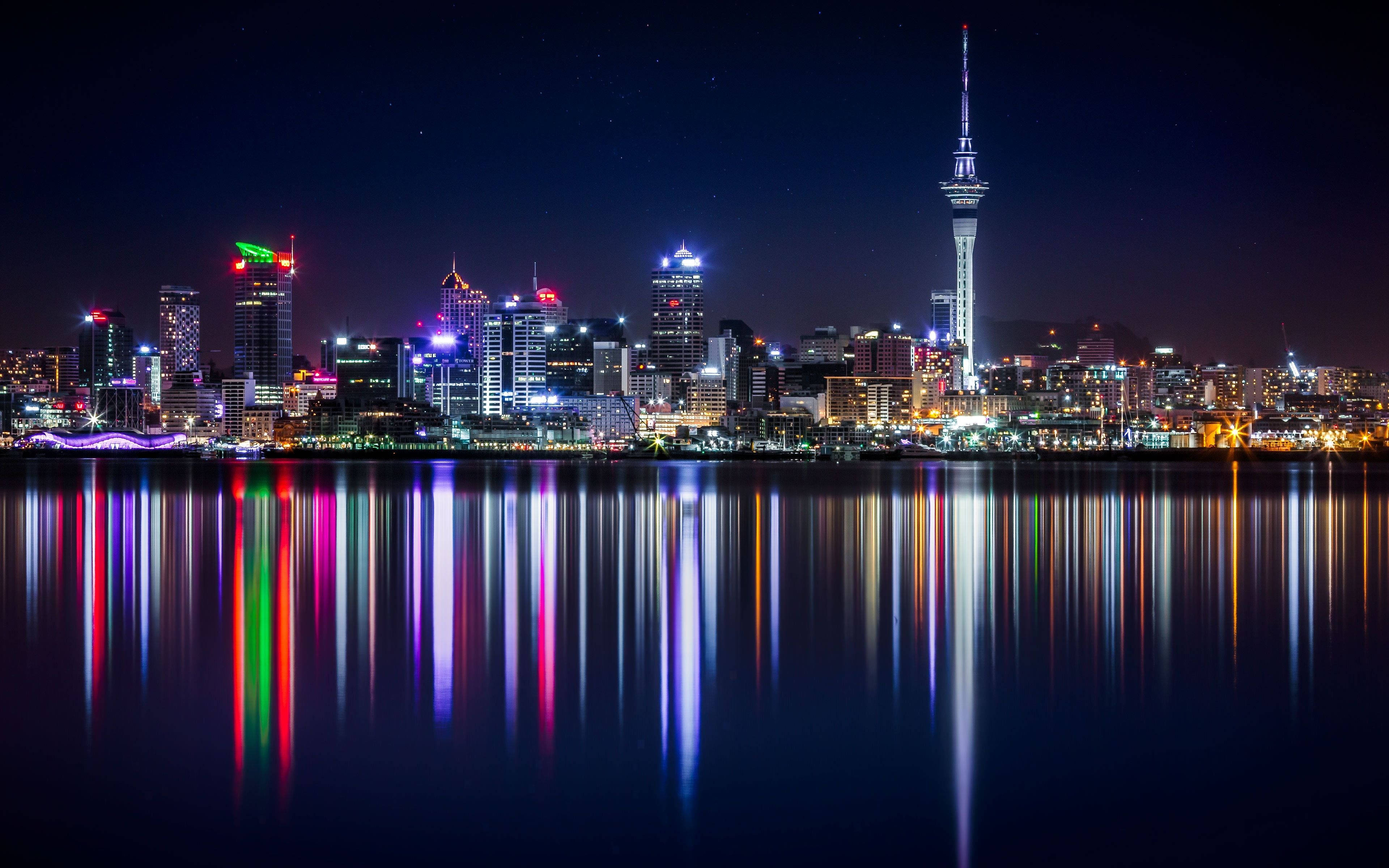 New 桌布 壁紙 — 充滿活力的城市天際線夜景，色彩繽紛的燈光在星空下平靜的水面上完美倒映。