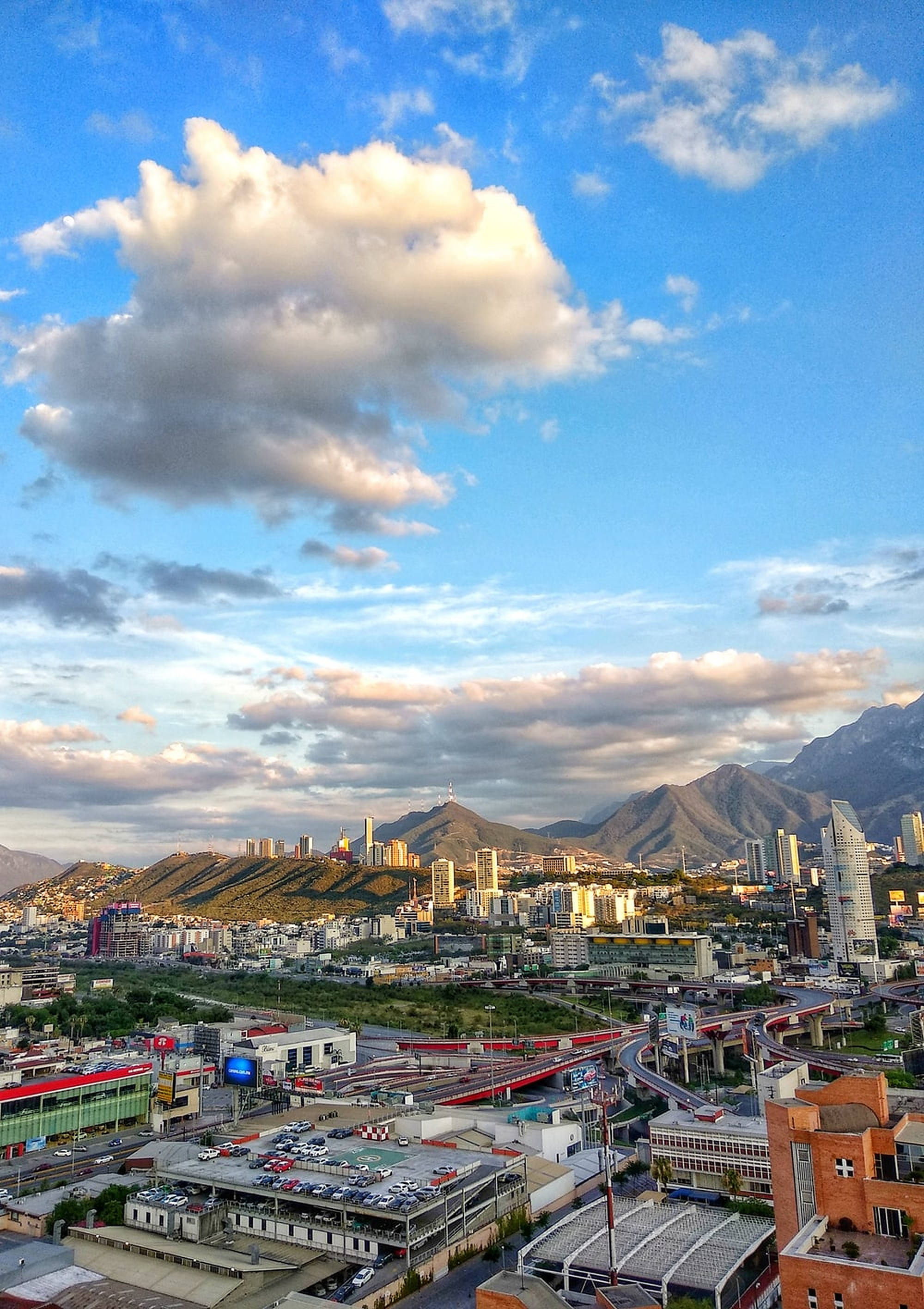 Monterrey 桌布 壁紙 — 充滿活力的城市景觀，坐落在群山之間，天空戲劇性變幻，雲朵蓬鬆，夕陽金光閃耀。