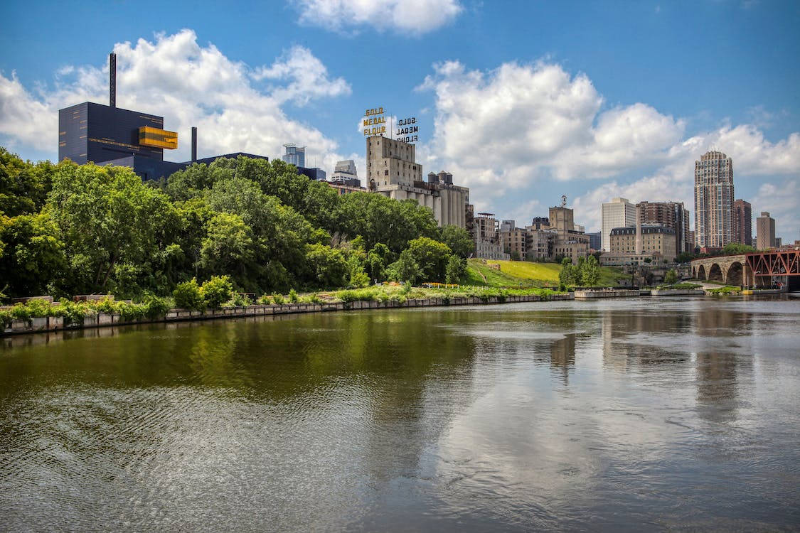 城市 桌布 壁紙 — 充滿活力的城市景觀，河流倒映著摩天大樓和茂密的綠樹，天空湛藍，白雲朵朵。