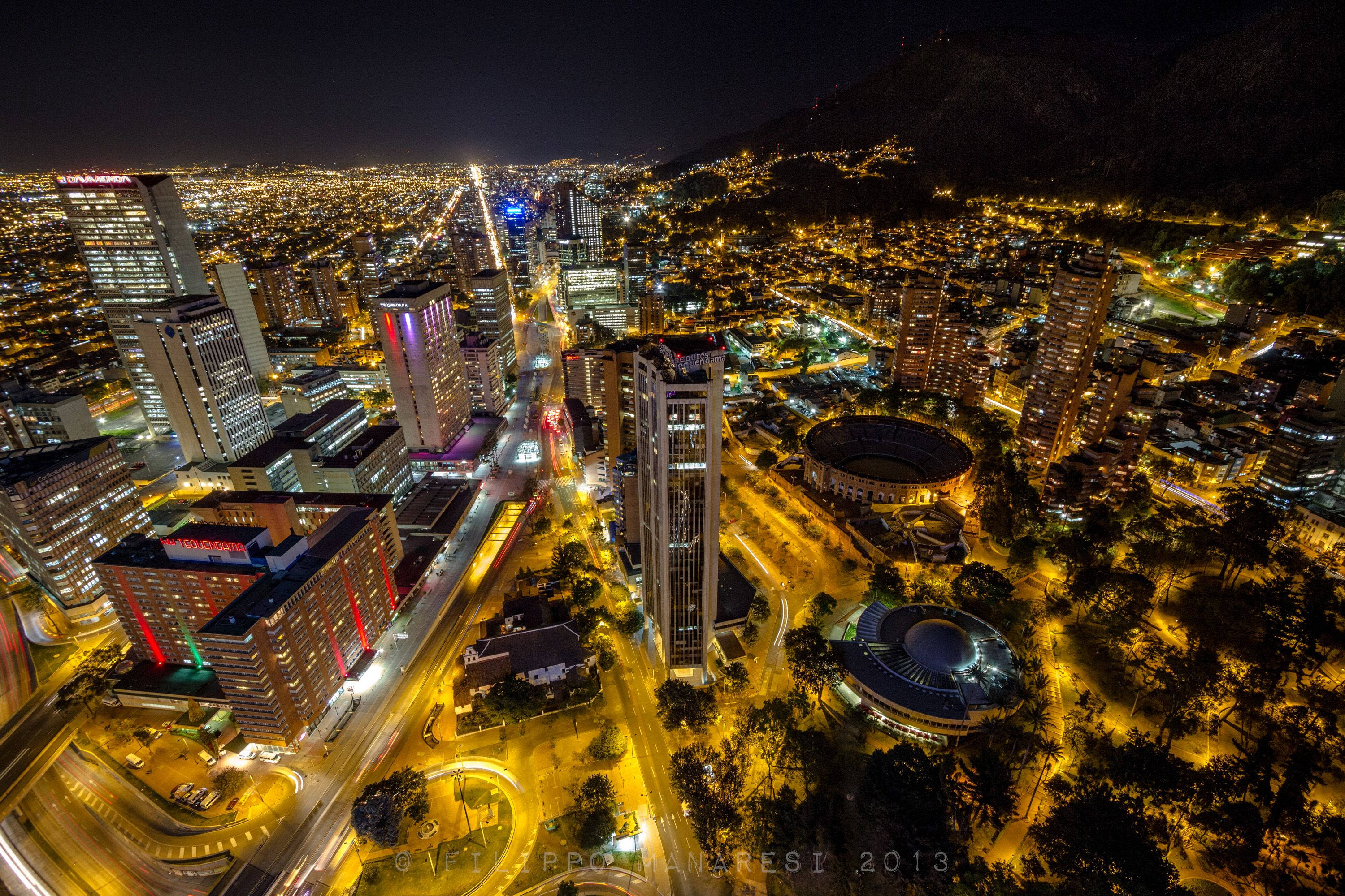 Bogota 桌布 壁紙 — 充滿活力的城市景觀夜間鳥瞰圖，閃爍的摩天大樓、蜿蜒的道路和山脈背景。