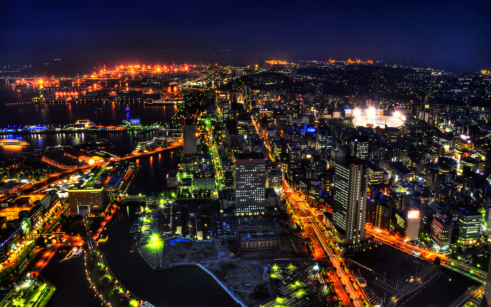 城市 桌布 壁紙 — 充滿活力的城市景觀夜間鳥瞰圖，閃爍的摩天大樓、河畔橋樑和燈火通明的街道倒映在水中。
