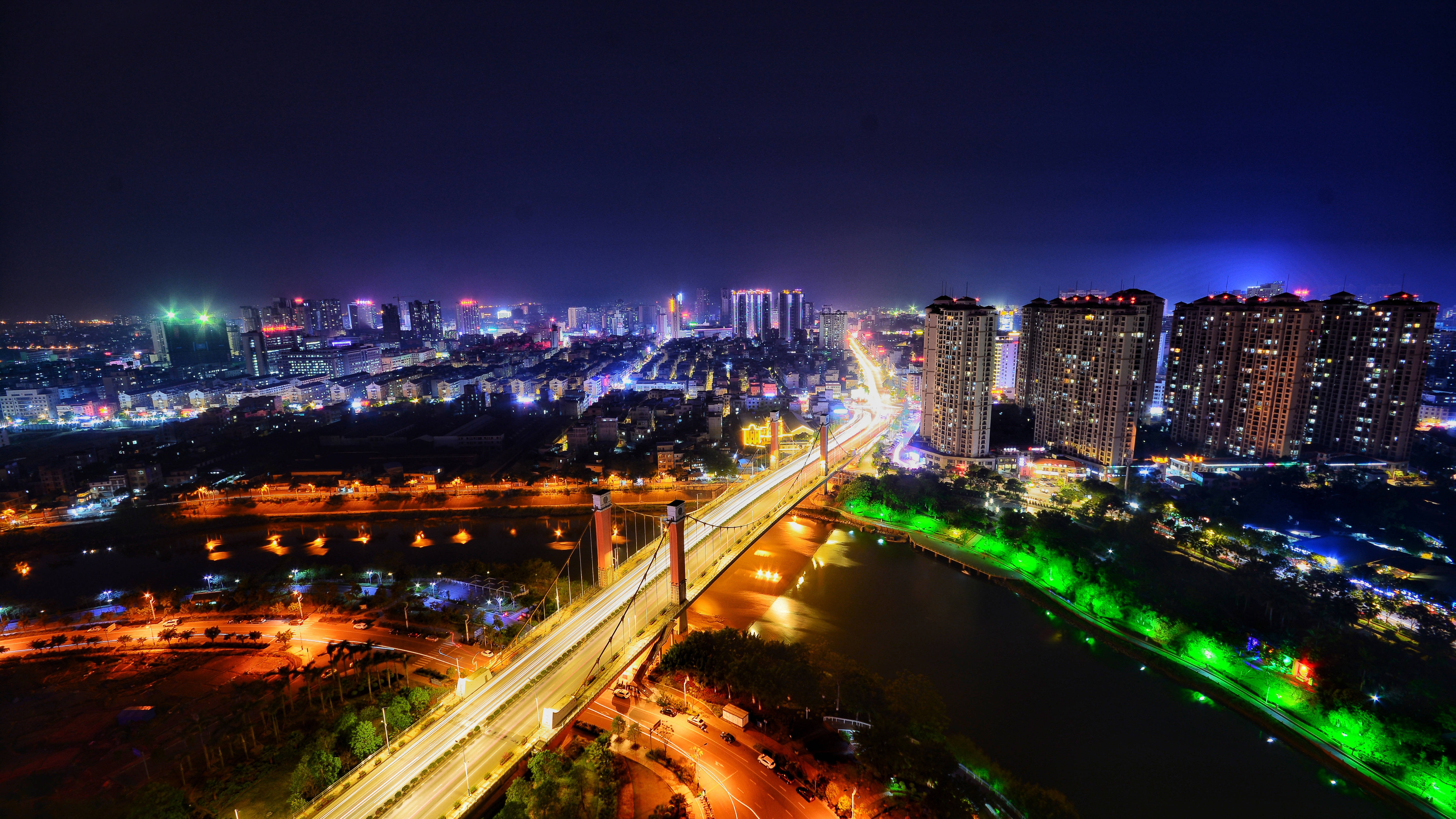 Dark 4K 桌布 壁紙 — 充滿活力的城市景觀的夜間鳥瞰圖，有發光的摩天大樓、一座橫跨河流的燈光橋樑和車流的燈光軌跡。