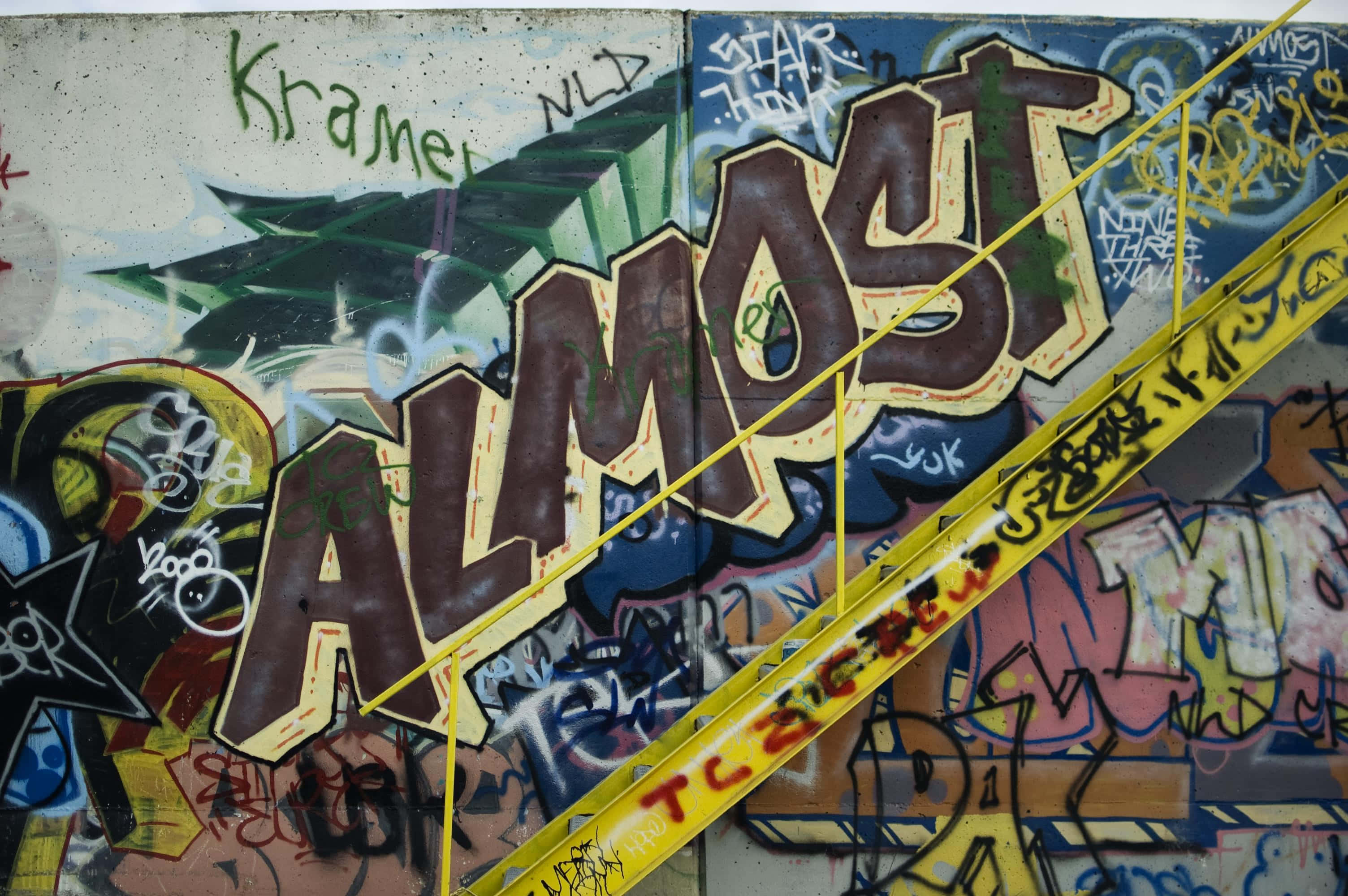 Graffiti Art 桌布 壁紙 — 充滿活力的塗鴉牆，帶有粗體 'ALMOST' 字樣和黃色警告膠帶斜穿過混亂的街頭藝術。
