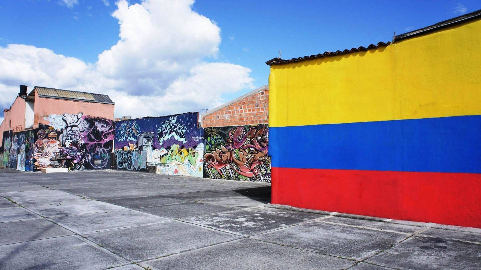 Colombia 桌布 壁紙 — 充滿活力的塗鴉牆壁和醒目的哥倫比亞國旗壁畫點綴著城市庭院，上方是晴朗的藍天和蓬鬆的白雲。