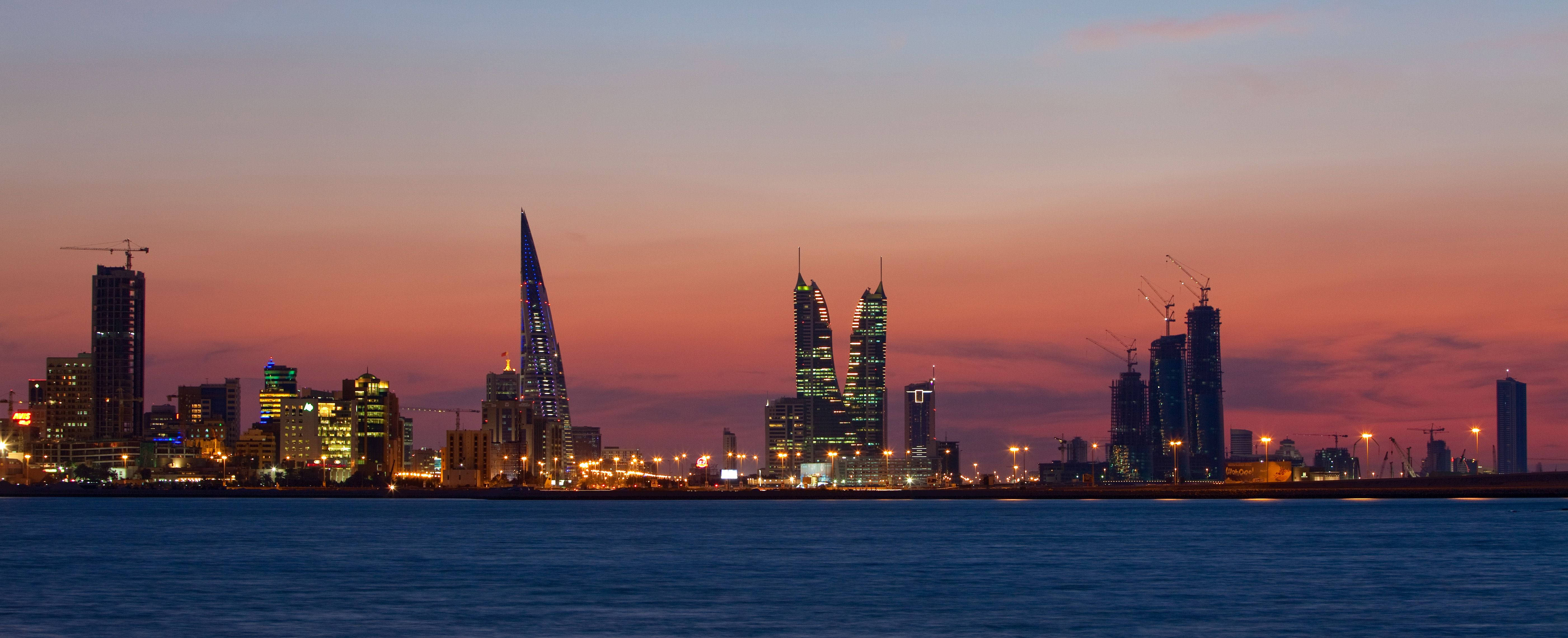 Bahrain 桌布 壁紙 — 充滿活力的夕陽將天空染成橙色和粉紅色，現代摩天大樓在平靜的水面上映襯著地平線閃耀。