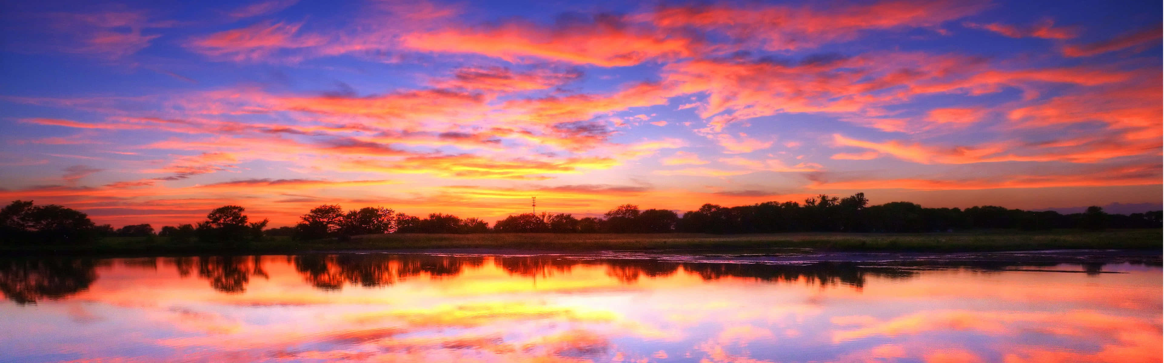 Panoramic Desktop 桌布 壁紙 — 充滿活力的夕陽將天空染成火熱的橙色和紫色，完美地倒映在平靜的湖面上，地平線上的樹木形成剪影。