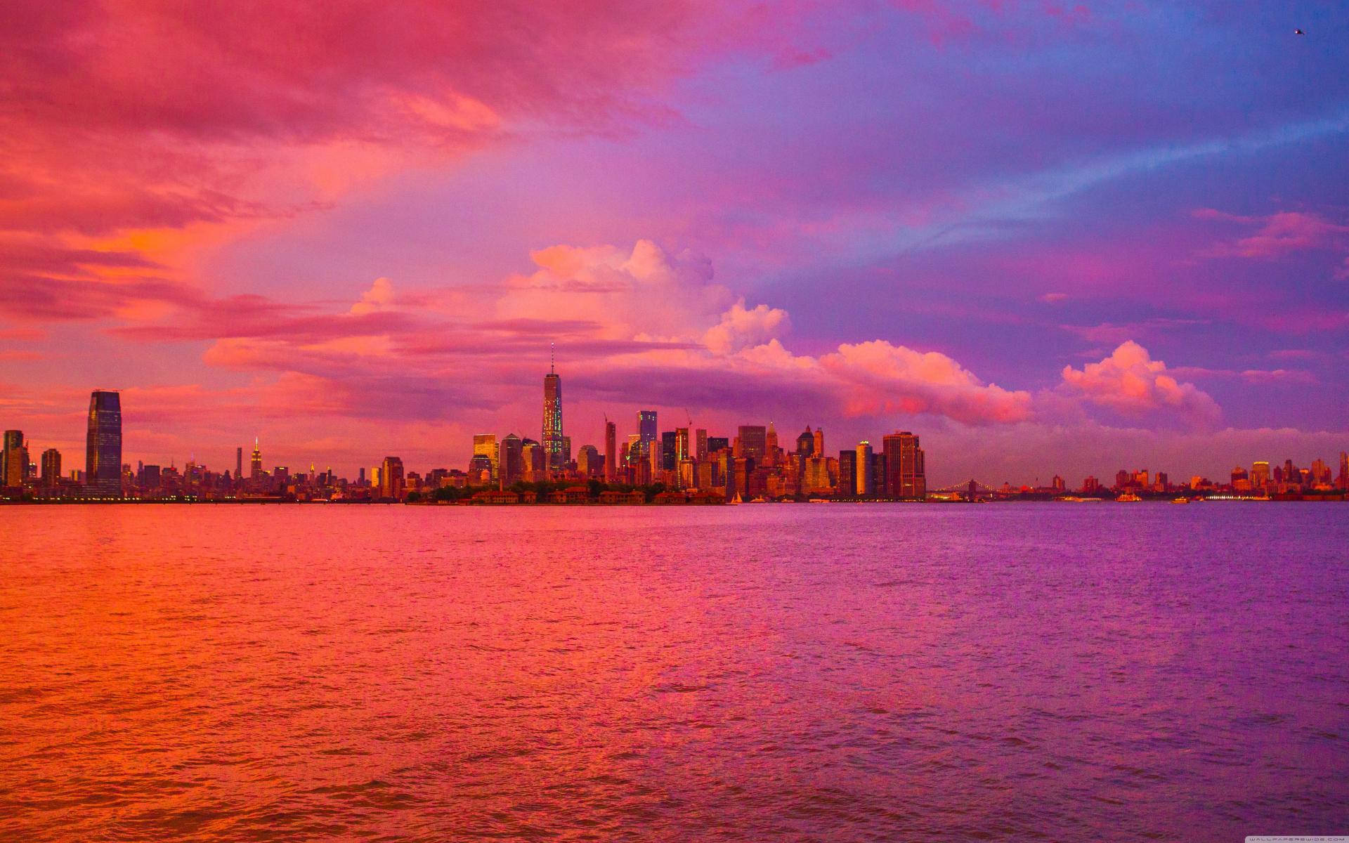 雲朵 桌布 壁紙 — 充滿活力的夕陽將廣闊的城市天際線沐浴在火熱的粉紅色和紫色色調中，倒映在平靜的水面上。