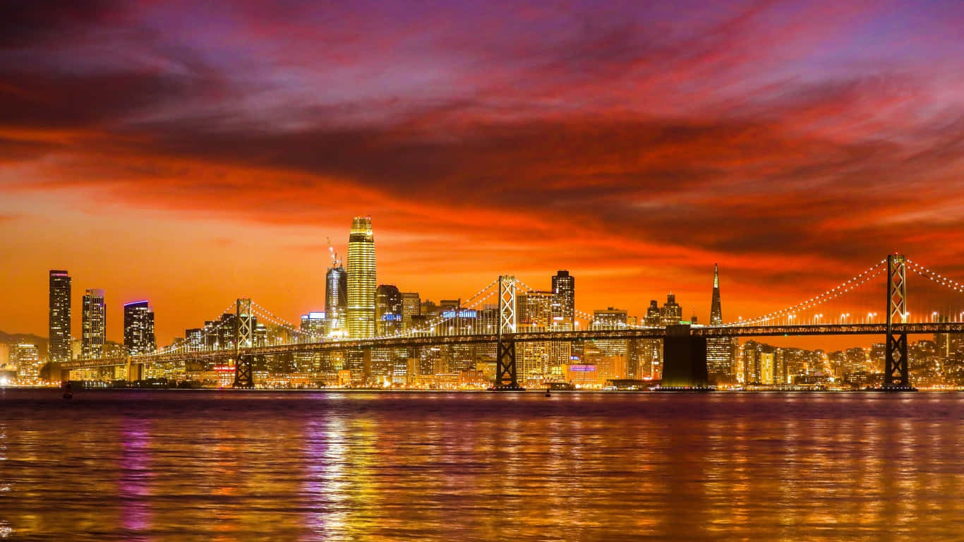 1366x768 San Francisco 桌布 壁紙 — 充滿活力的夕陽用火紅和橙色渲染天空，映照著發光的城市天際線和一座亮燈的吊橋，倒映在平靜的水面上。