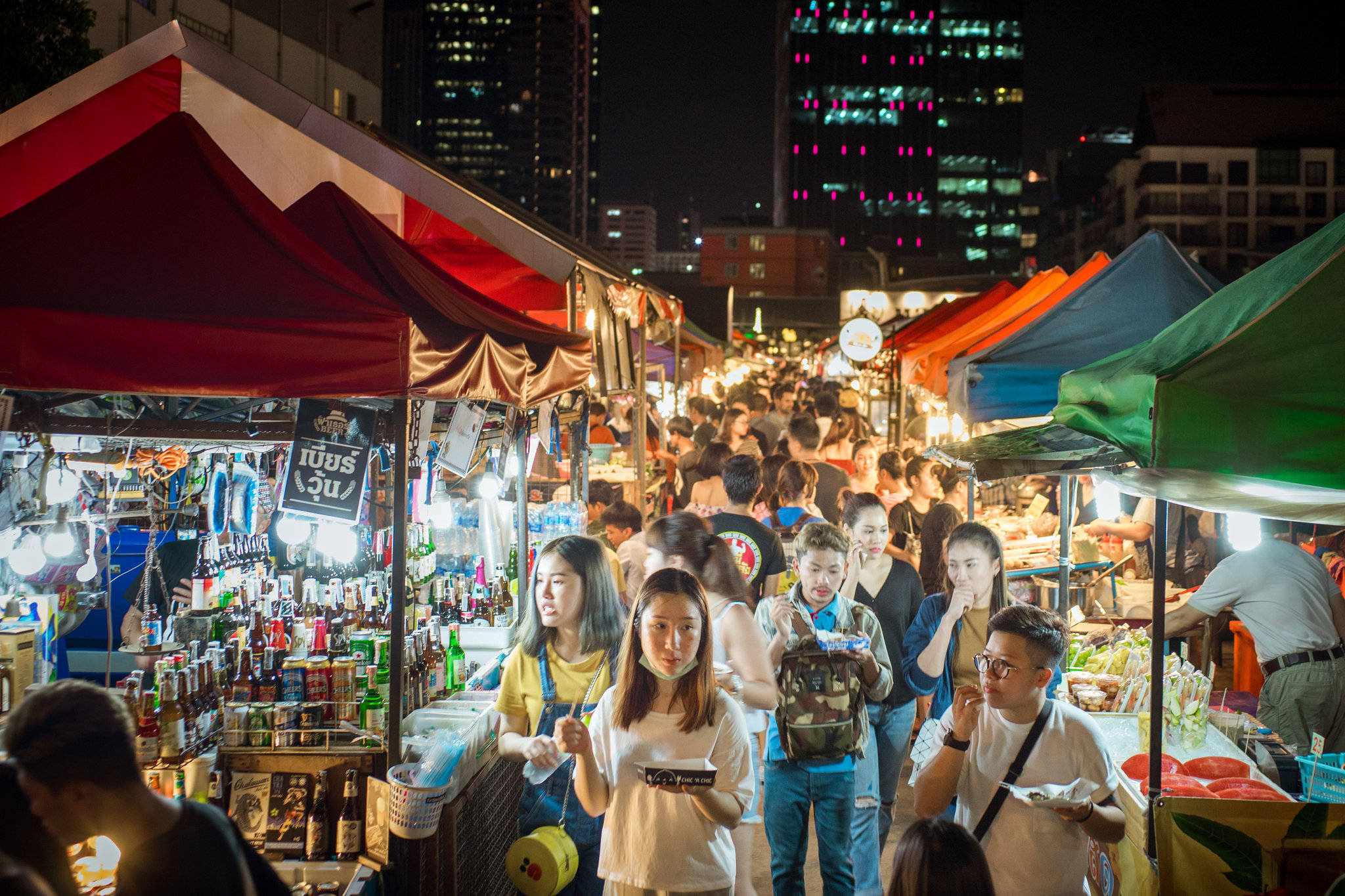 Bangkok 桌布 壁紙 — 充滿活力的夜市，色彩繽紛的帳篷下擠滿了人，溫暖的燈籠照亮了背景中燈火通明的摩天大樓。