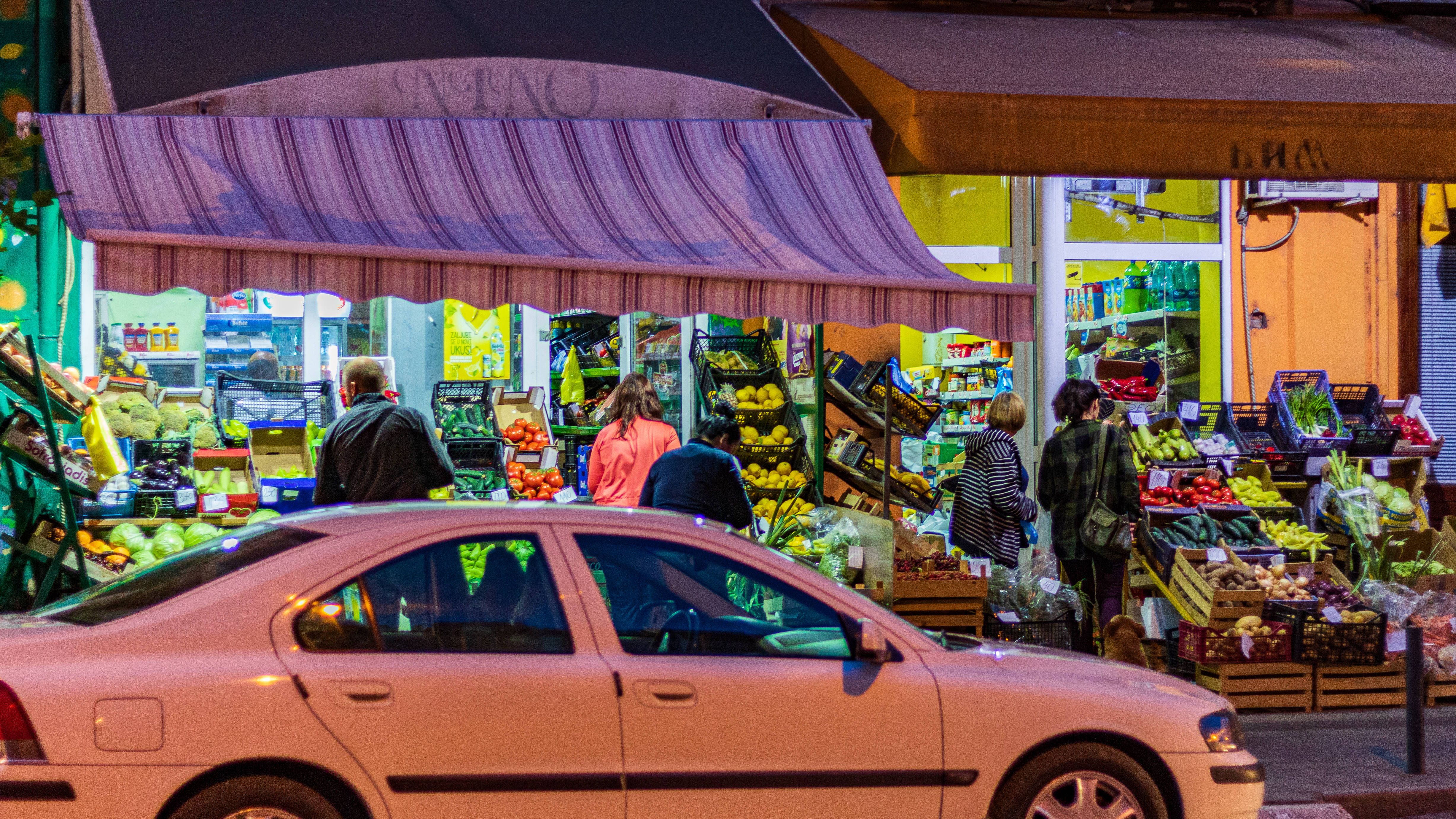 Serbia 桌布 壁紙 — 充滿活力的夜市攤位，在閃爍燈光下擺滿色彩繽紛的農產品，購物者瀏覽，一輛粉紅色汽車駛過。
