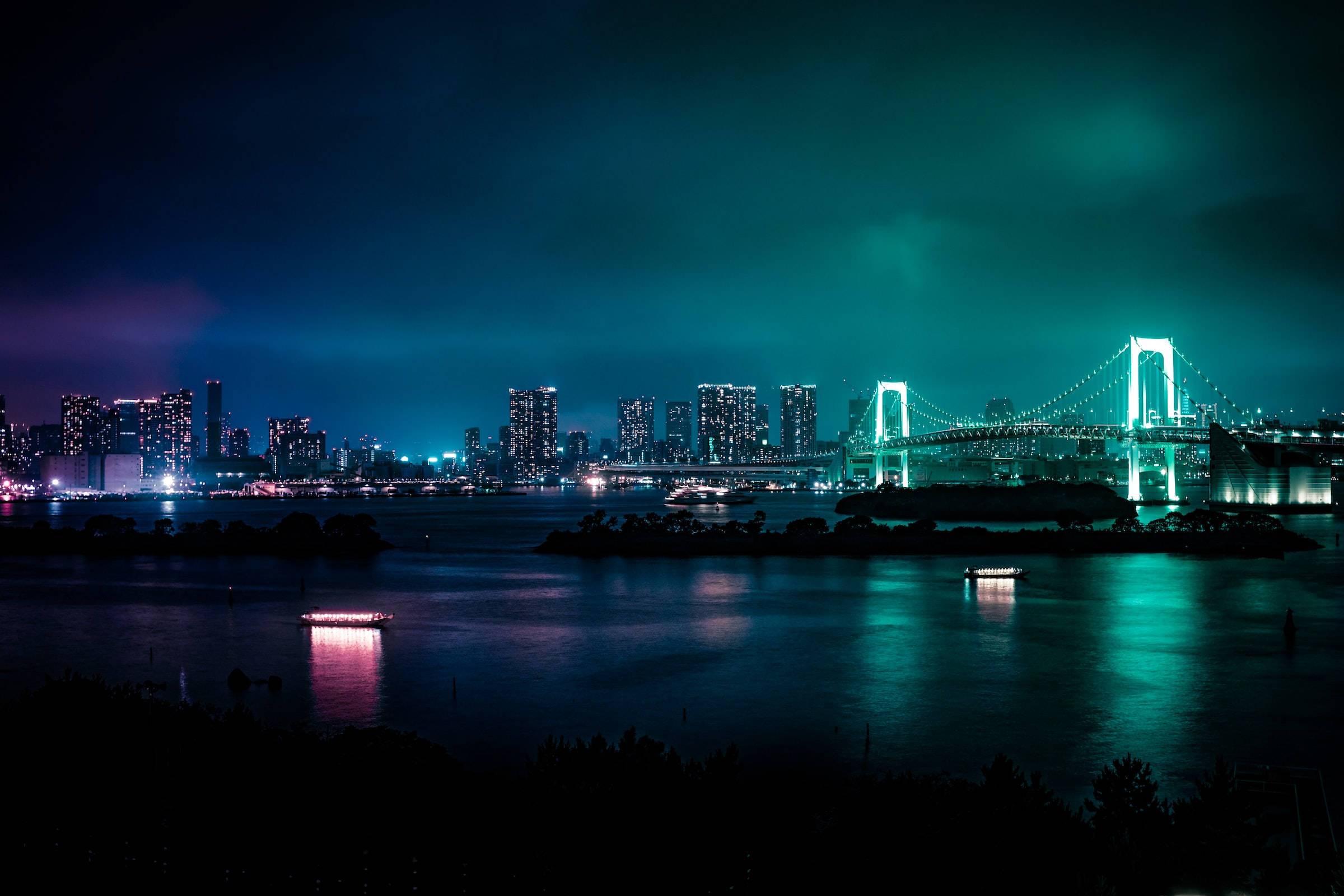 夜空 桌布 壁紙 — 充滿活力的夜景，以發光的彩虹橋橫跨水面，倒影和遠處摩天大樓在深藍綠色天空下。