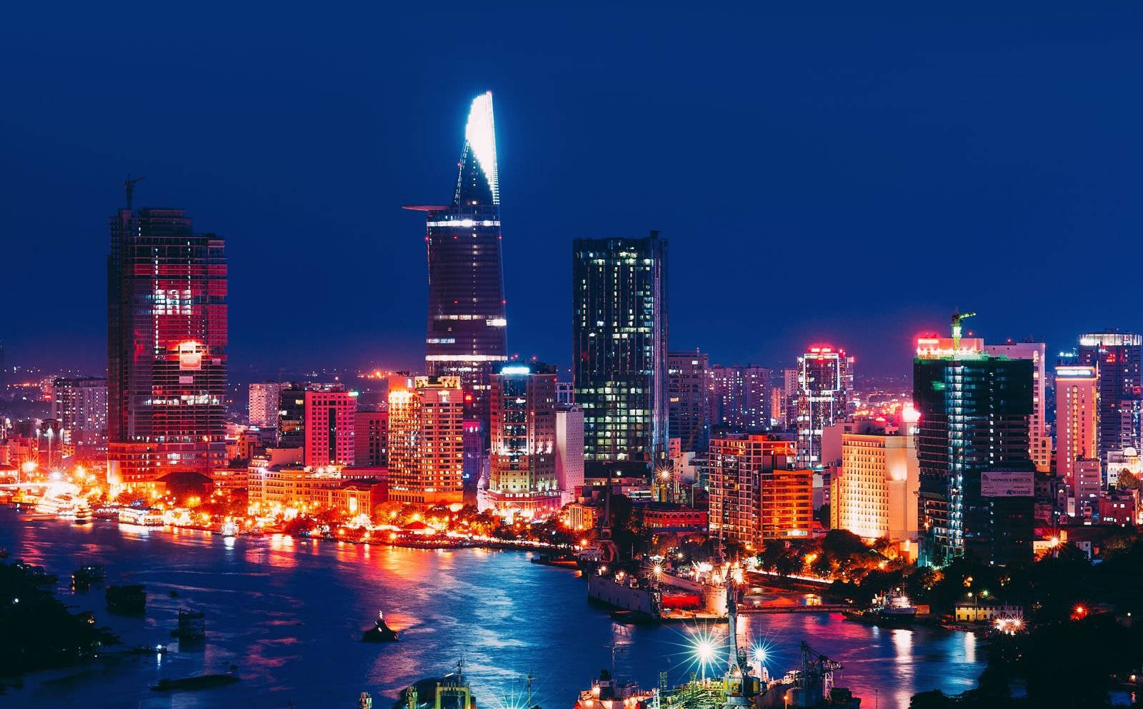 胡志明市 桌布 壁紙 — 充滿活力的夜景，在深藍色天空下，發光的摩天大樓倒映在閃爍的河流上。