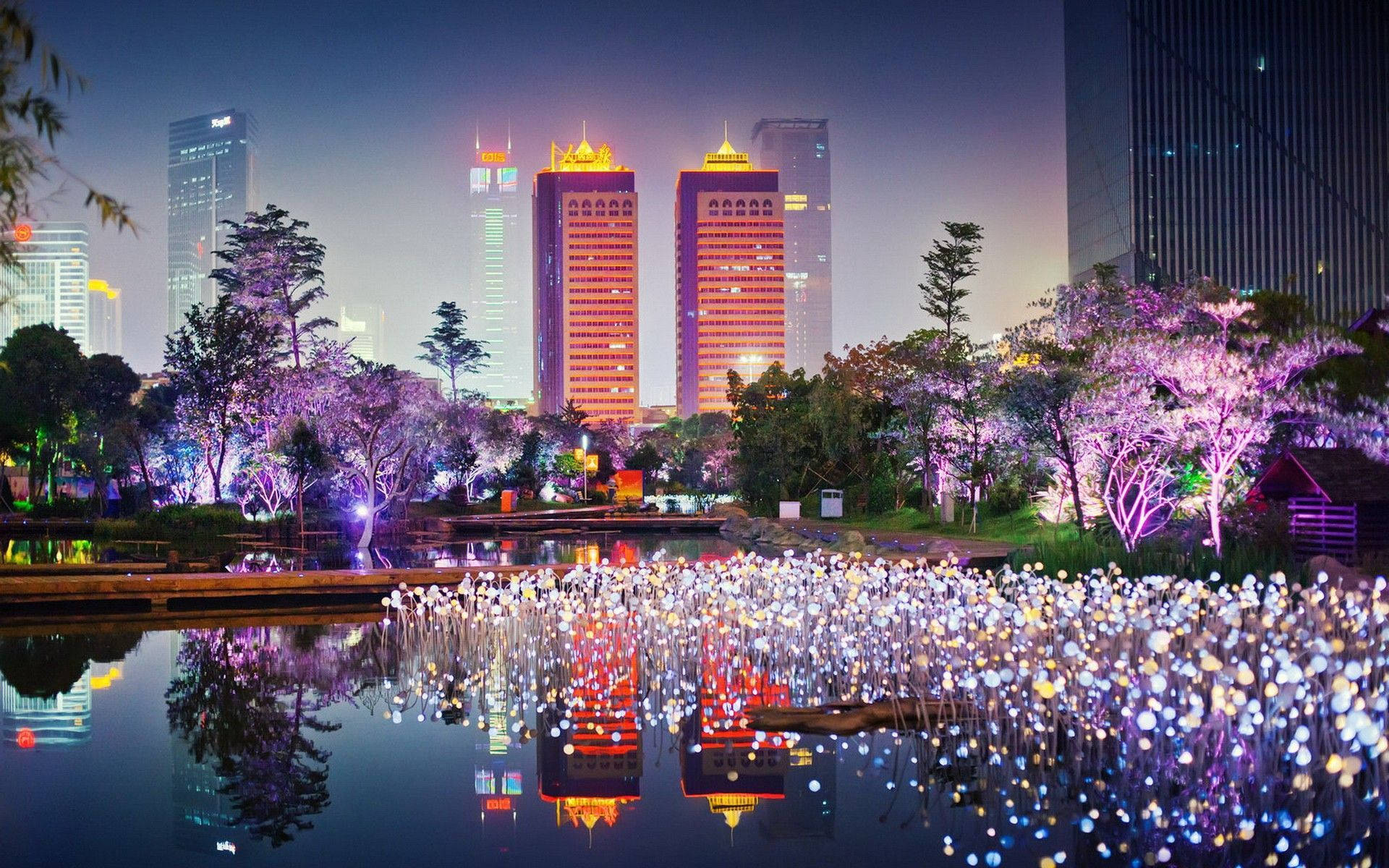 lights 桌布 壁紙 — 充滿活力的夜景，有發光的紫色樹木和閃爍的燈光，倒映在平靜的池塘中，周圍環繞著現代摩天大樓。