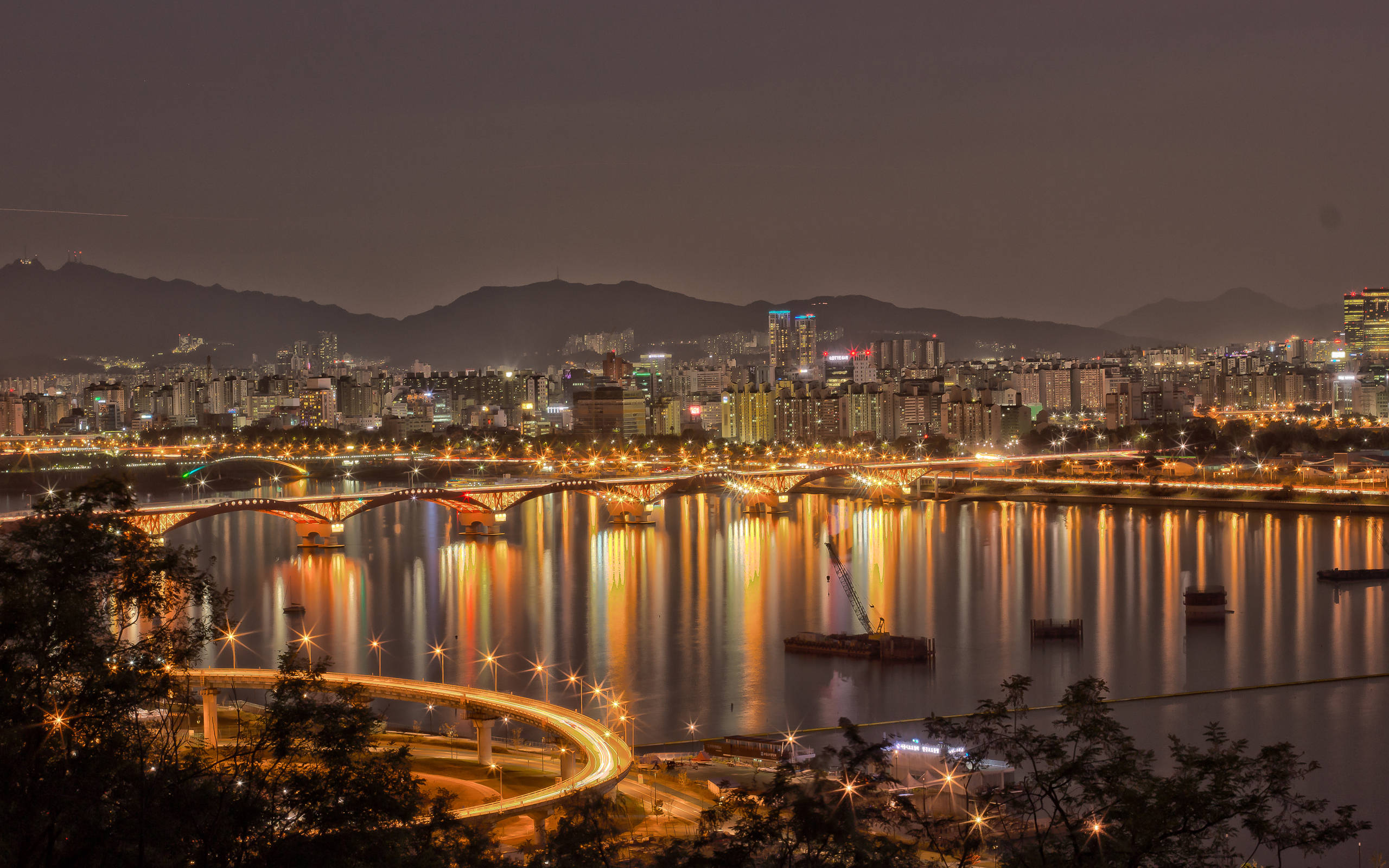 南韓 桌布 壁紙 — 充滿活力的夜景，發光的橋樑和摩天大樓倒映在平靜的河流中，周圍環繞著黑暗的山脈。