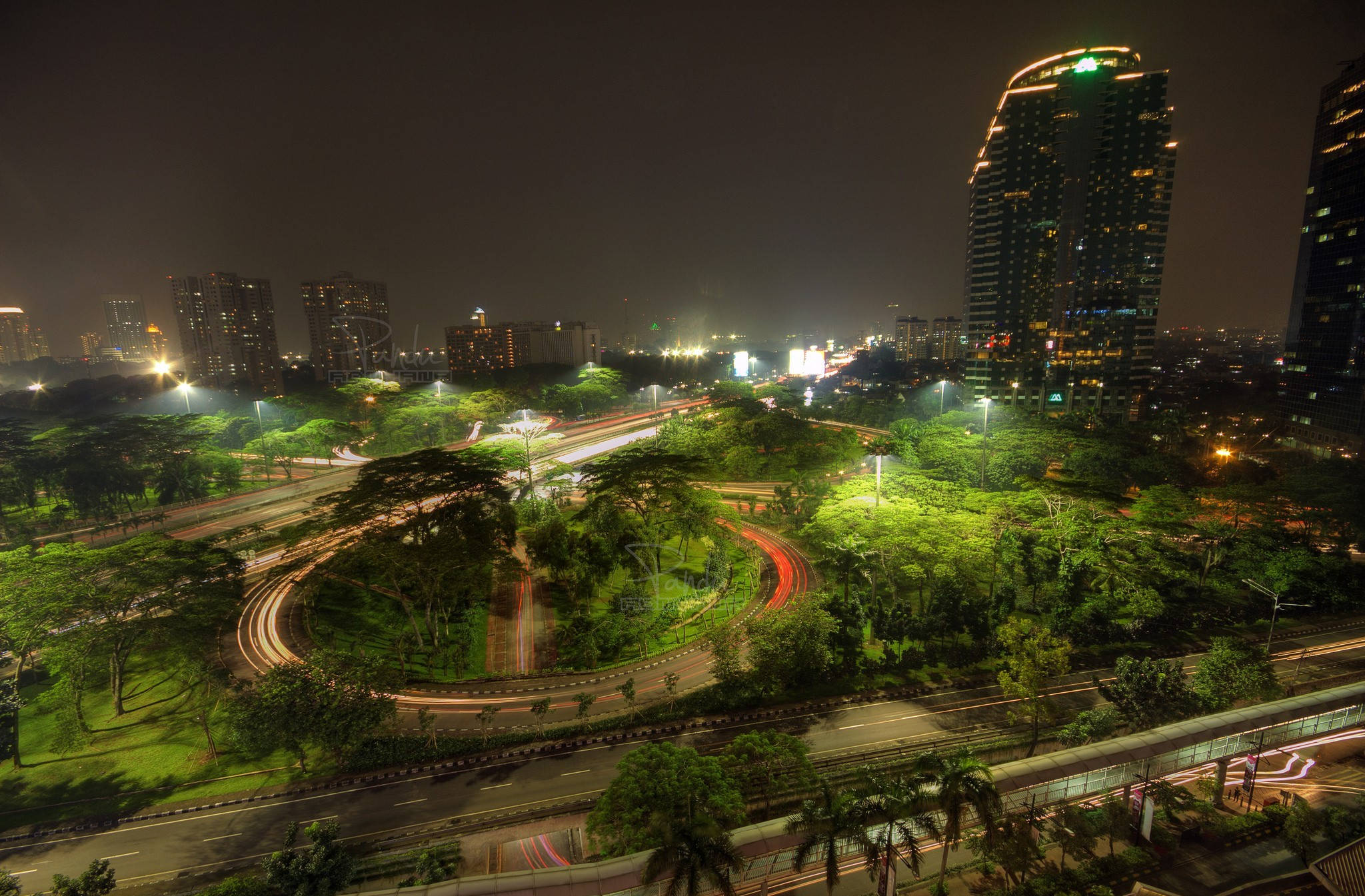 Jakarta 桌布 壁紙 — 充滿活力的夜景，閃爍的摩天大樓和蜿蜒穿過茂密綠色公園的汽車光跡。