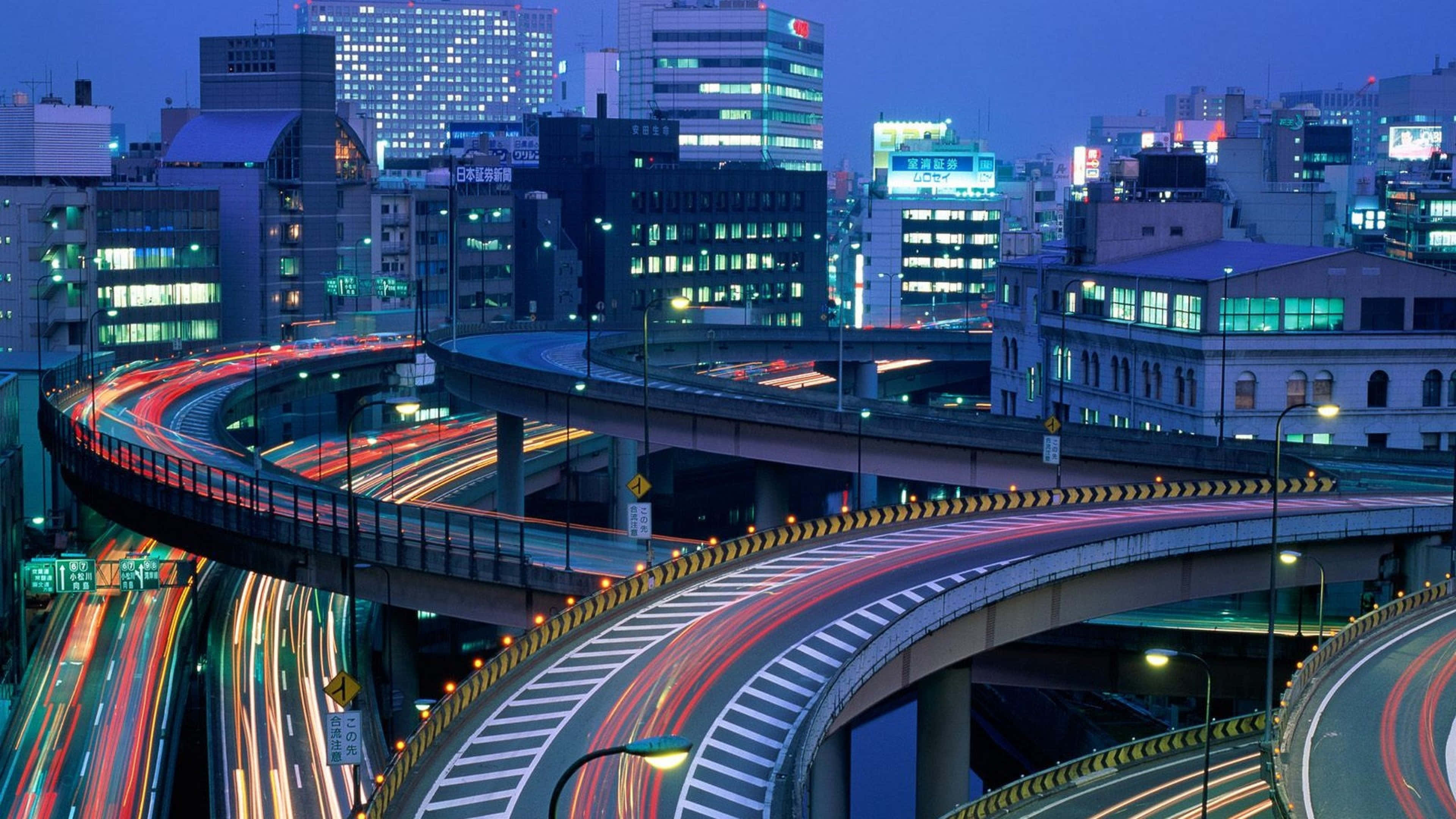 Infrastructure 桌布 壁紙 — 充滿活力的夜景，閃爍的摩天大樓和蜿蜒高架公路上車流留下的發光軌跡。
