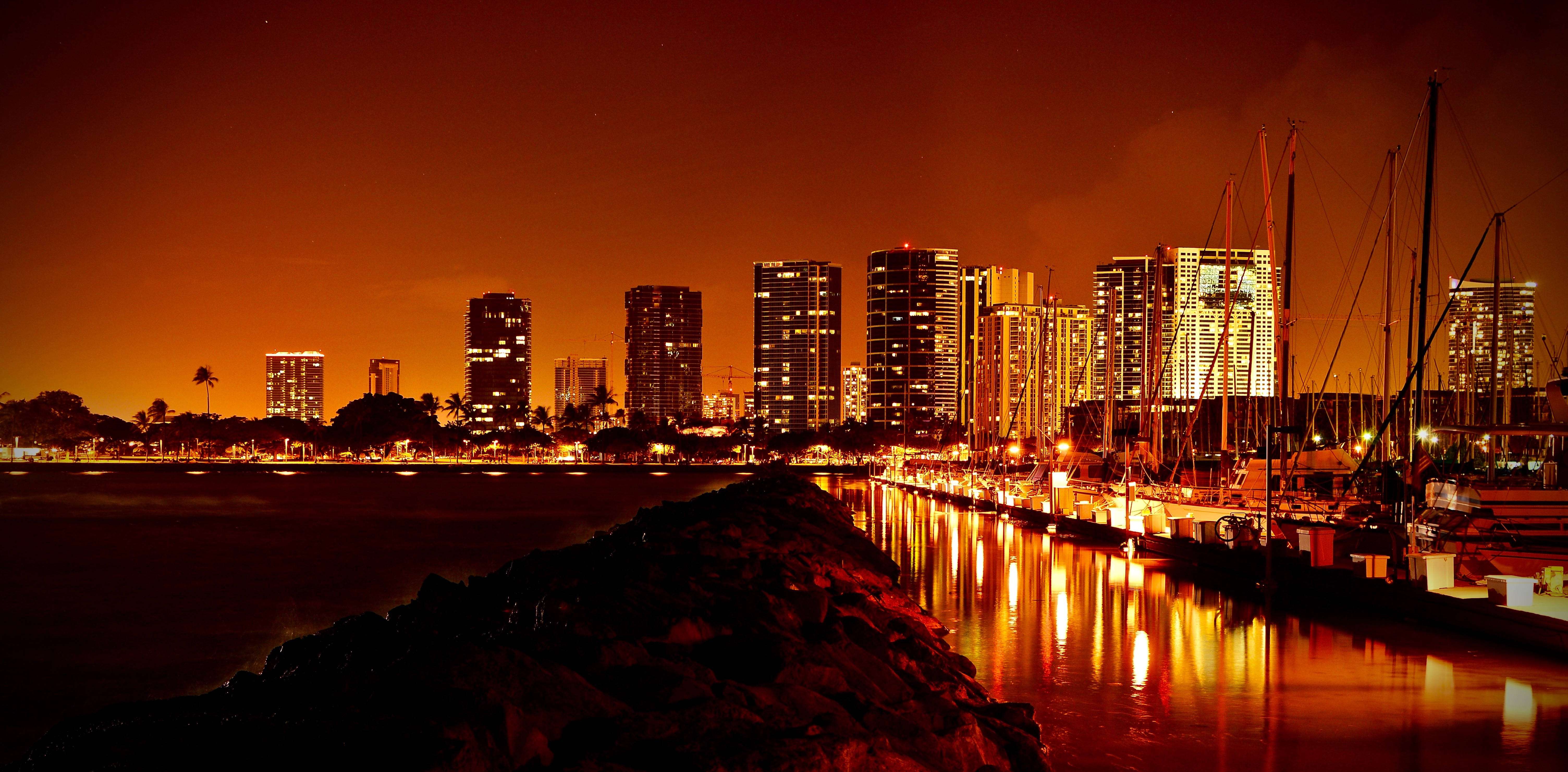夜晚 桌布 壁紙 — 充滿活力的夜景，閃爍的摩天大樓和停泊的船隻倒映在溫暖的橙色水面上。