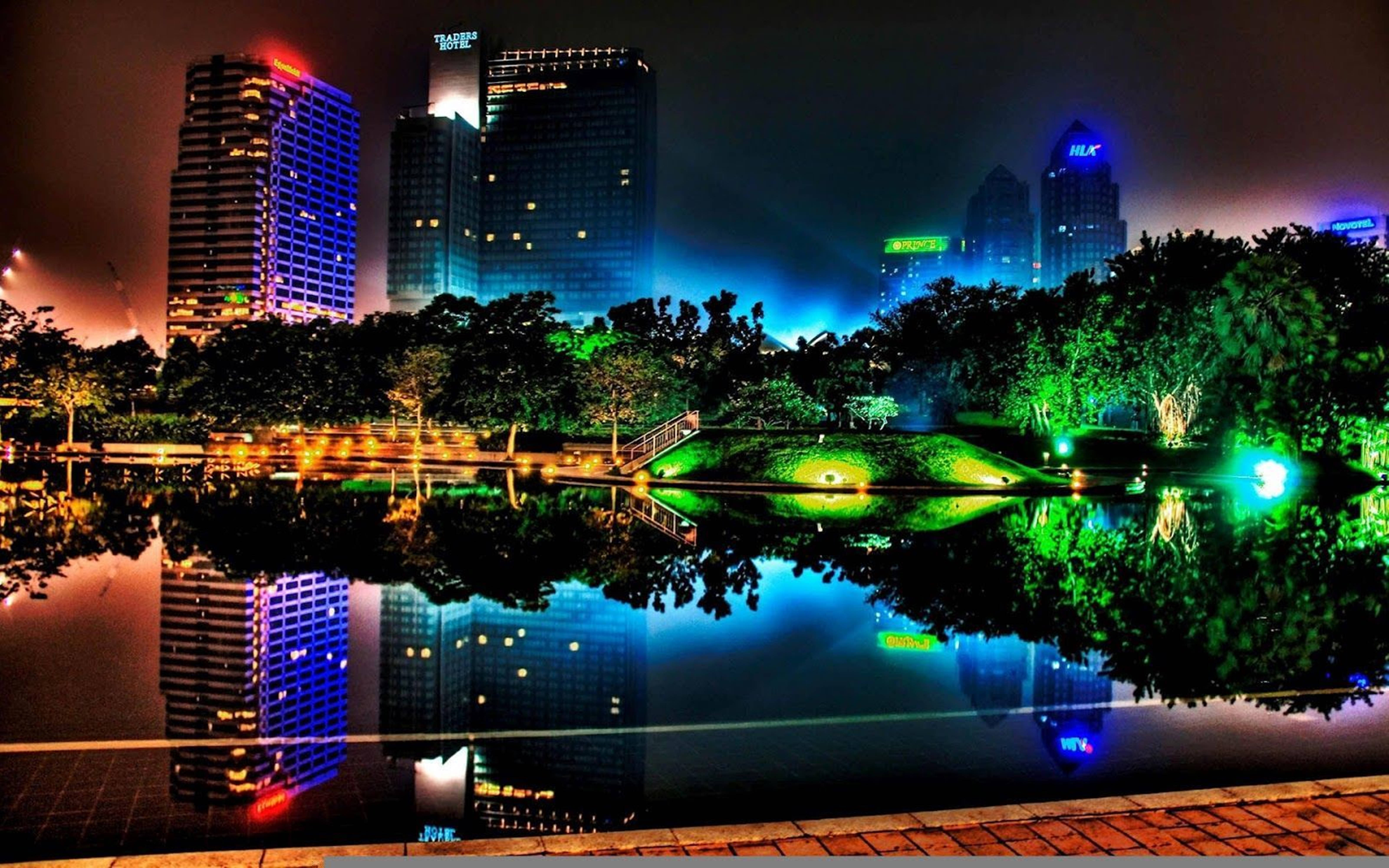 3D 桌布 壁紙 — 充滿活力的夜景，霓虹燈照亮的高樓大廈倒映在平靜的池塘中，周圍環繞著發光的綠色樹木。
