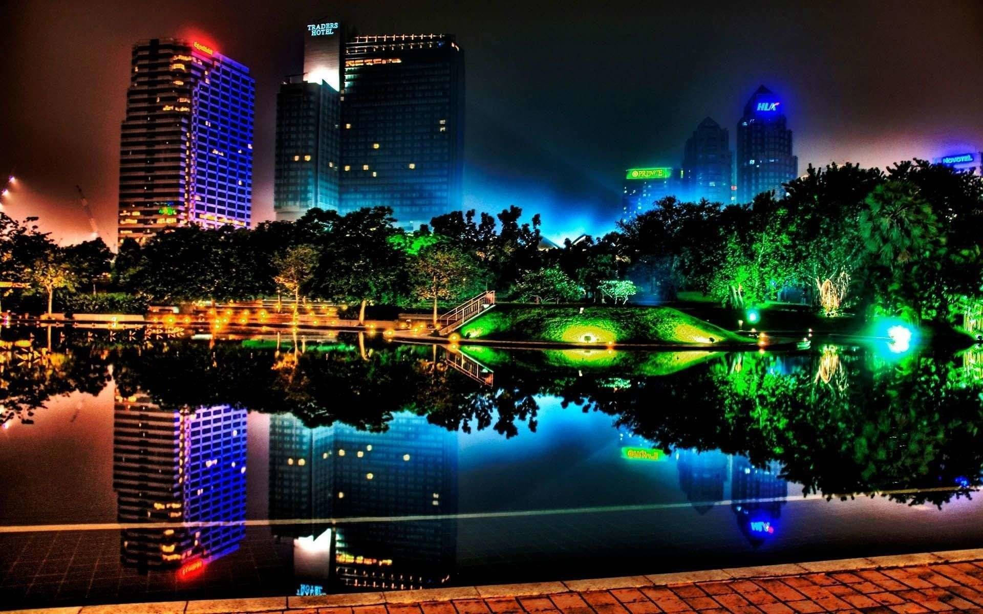 酷炫桌面 桌布 壁紙 — 充滿活力的夜景，霓虹燈閃爍的摩天大樓倒映在平靜的池塘中，周圍環繞著發光的綠色樹木。