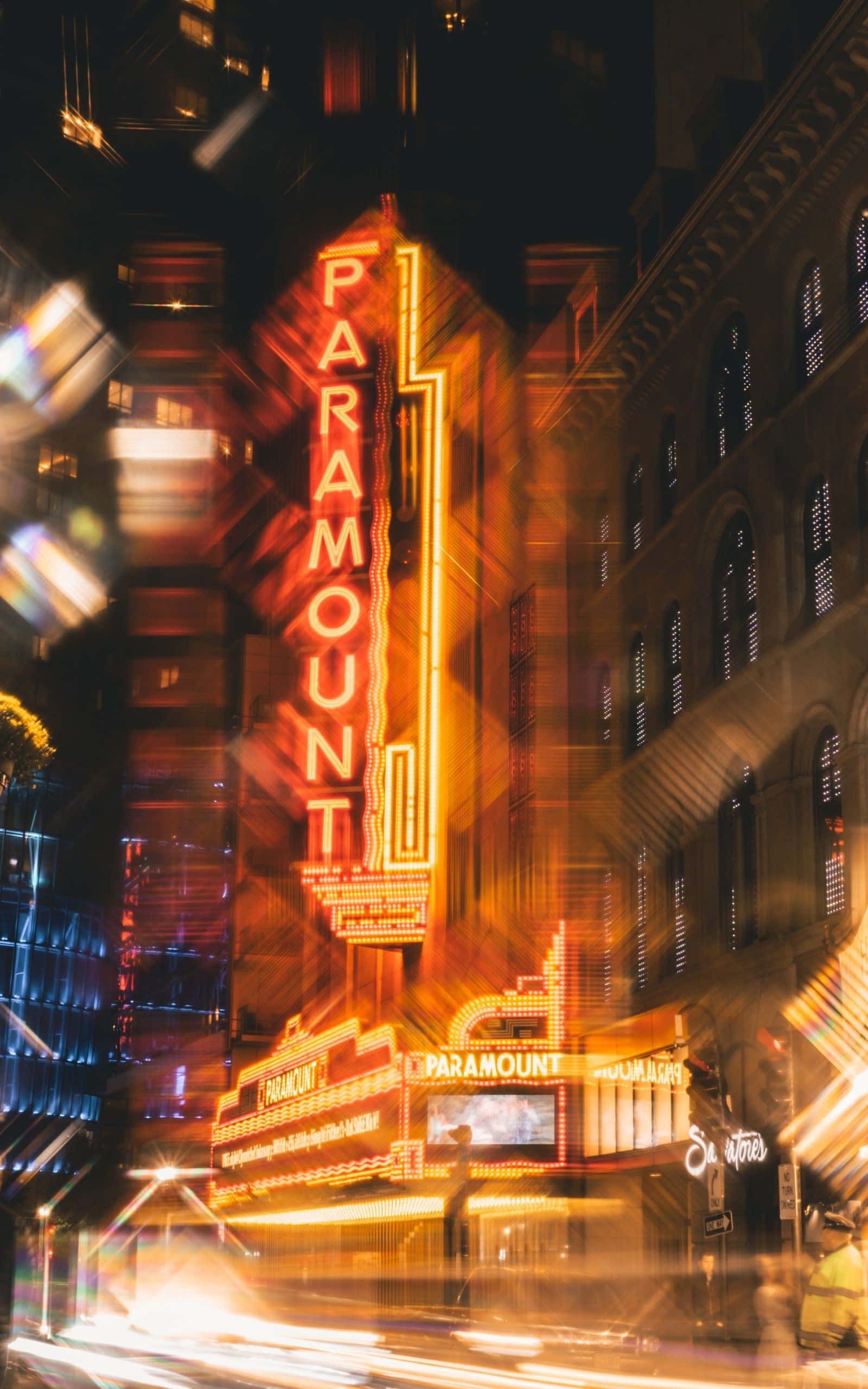 霓虹城市動態圖 桌布 壁紙 — 充滿活力的夜景城市景觀， featuring 發光的 Paramount 劇院標誌和來往車輛的光跡。