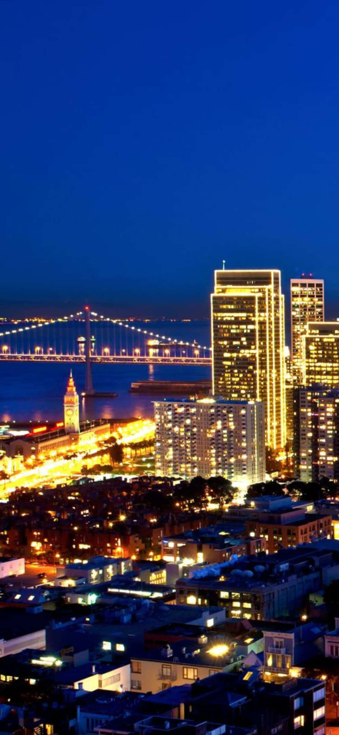 iPhone Xs California 桌布 壁紙 — 充滿活力的夜景城市景觀， featuring 發光的摩天大樓和一條橫跨黑暗水道的明亮照明吊橋。